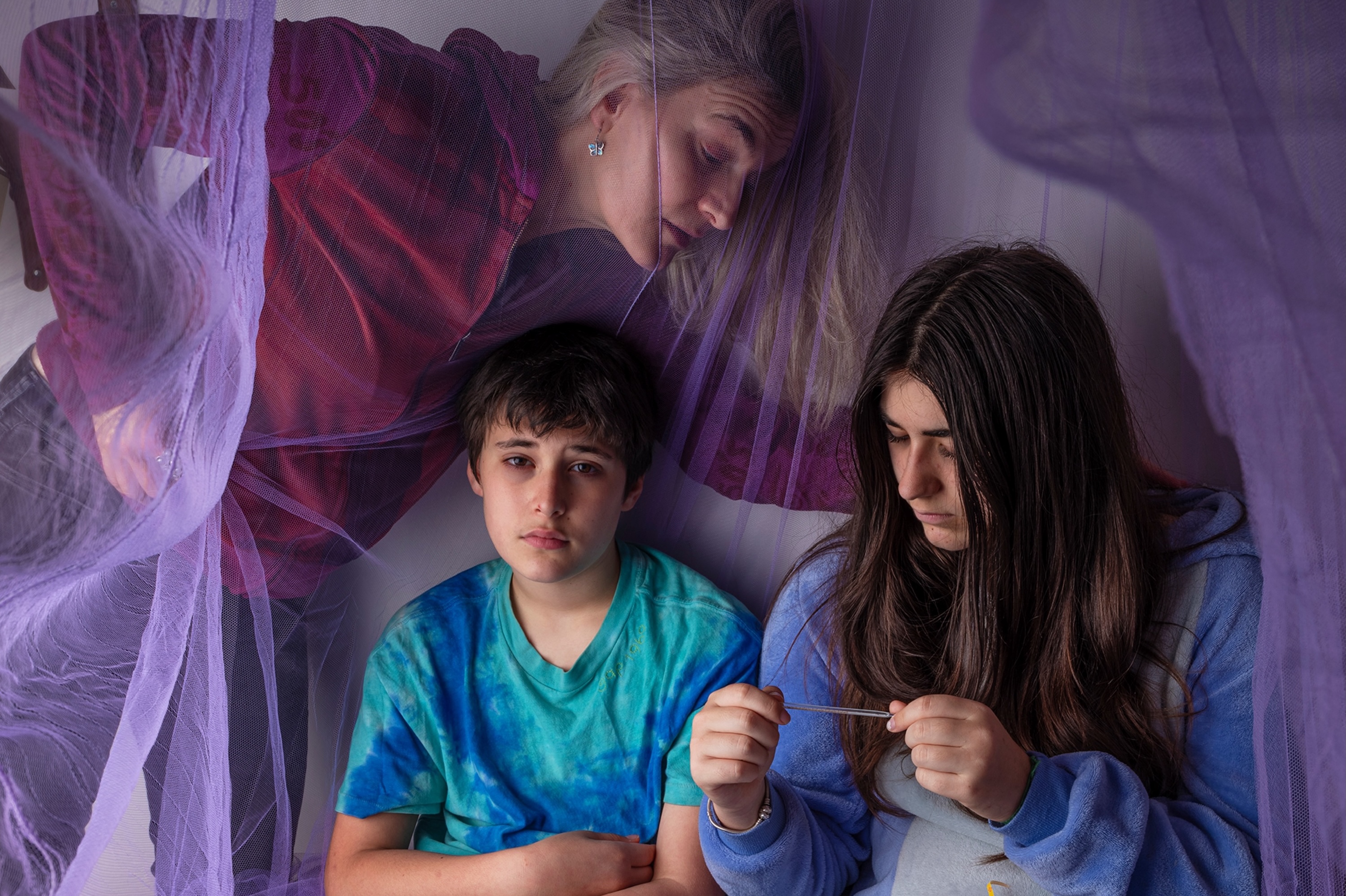 A woman checks the thermometer of her two children who sit inside a purple mosquito net