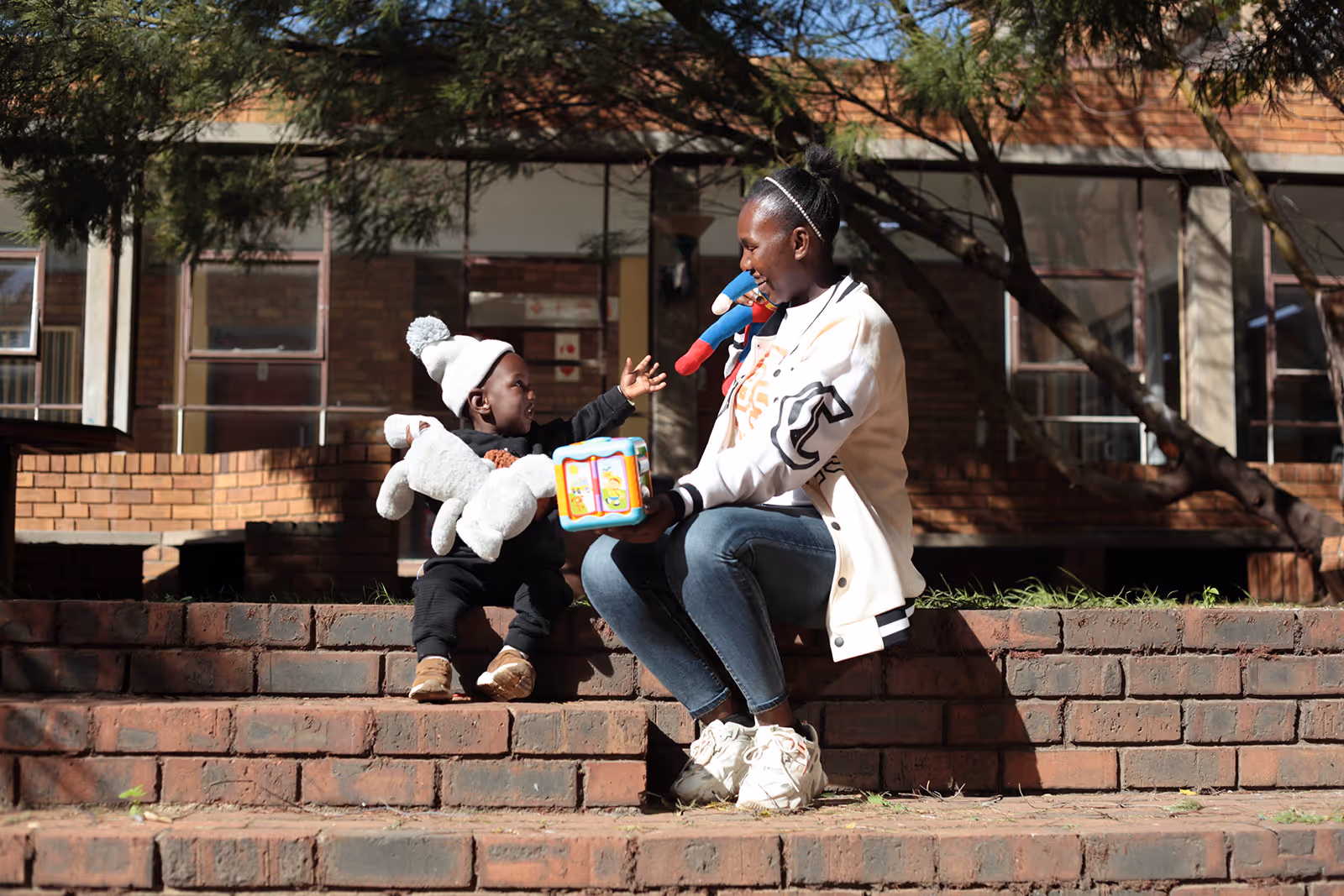 With buildings in the background, a woman wearing a whiter sweater and her young son, who’s holding a stuffed toy and wearing a winter hat, talk while sitting on steps.