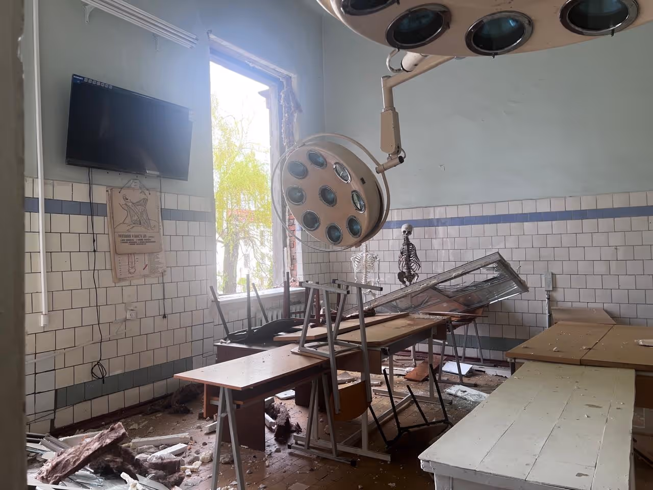 A destroyed classroom is filled with desks, chairs, and debris. A television still hangs on a wall to the left, and a skeleton meant for learning stands in the back of the room.
