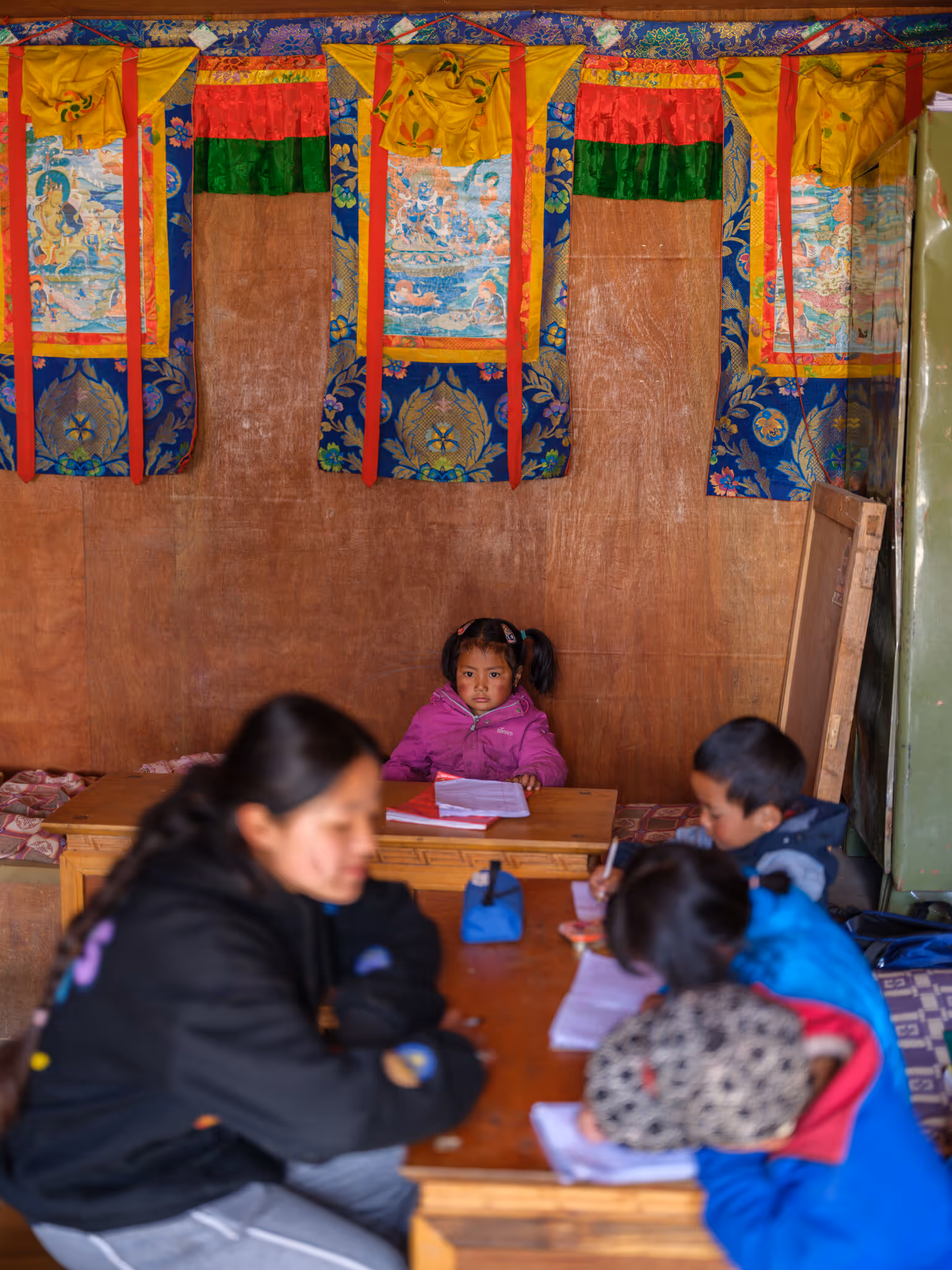 A small girl sits in a wooden makeshift classroom, wearing a bright pink winter jacket. Colorful traditional wall hangings decorate the wall behind her. Her young classmates study in the foreground. 