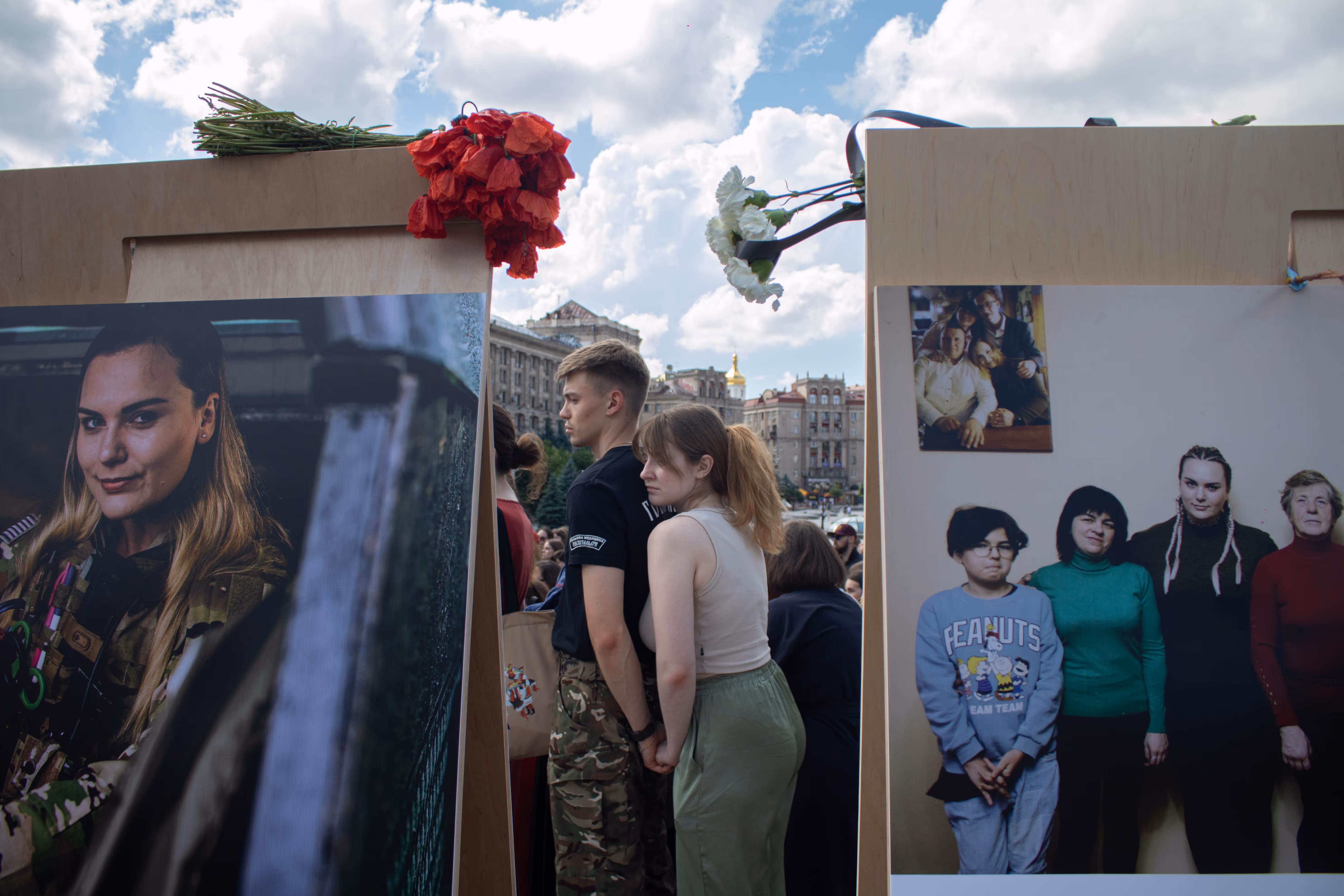 Mourners stand at a memorial service. In the foreground are two mounted photographs—on the left, a portrait of a woman, and on the right, a family. Red and white flowers are resting on top of the photographs. Behind the images two young people stand holding hands.