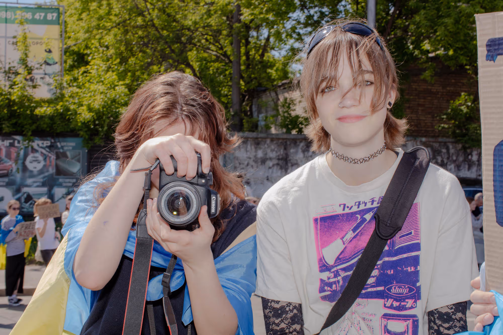 Two teenagers stand in a park facing the camera. On the left, one of them holds a camera in front of her face. The person on the right is making direct eye contact, wearing a graphic T-shirt, a necklace, and has sunglasses on top of her head.