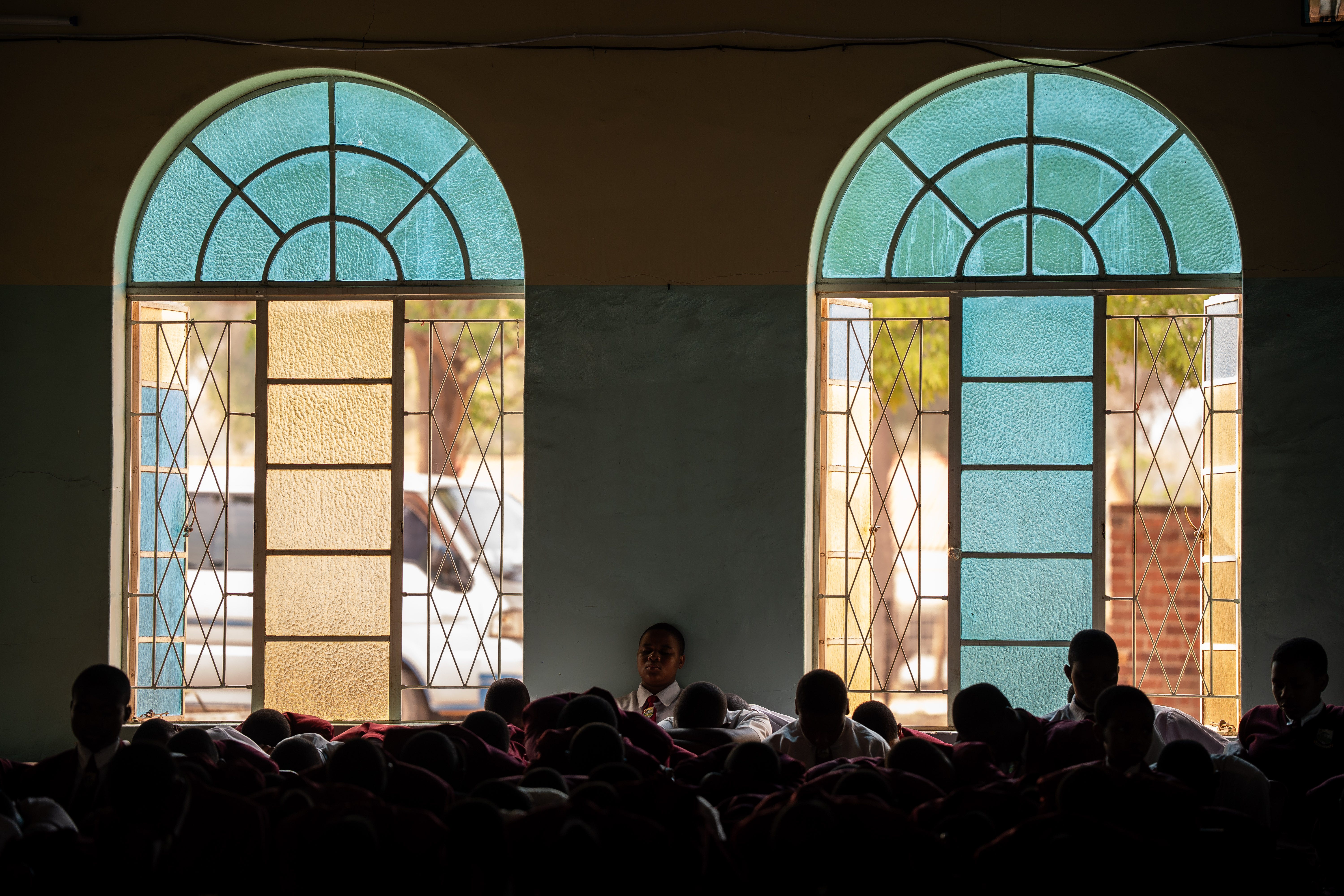 Students sit closely together inside a dim classroom, framed by two tall arched windows with blue and yellow glass panes that let in daylight.
