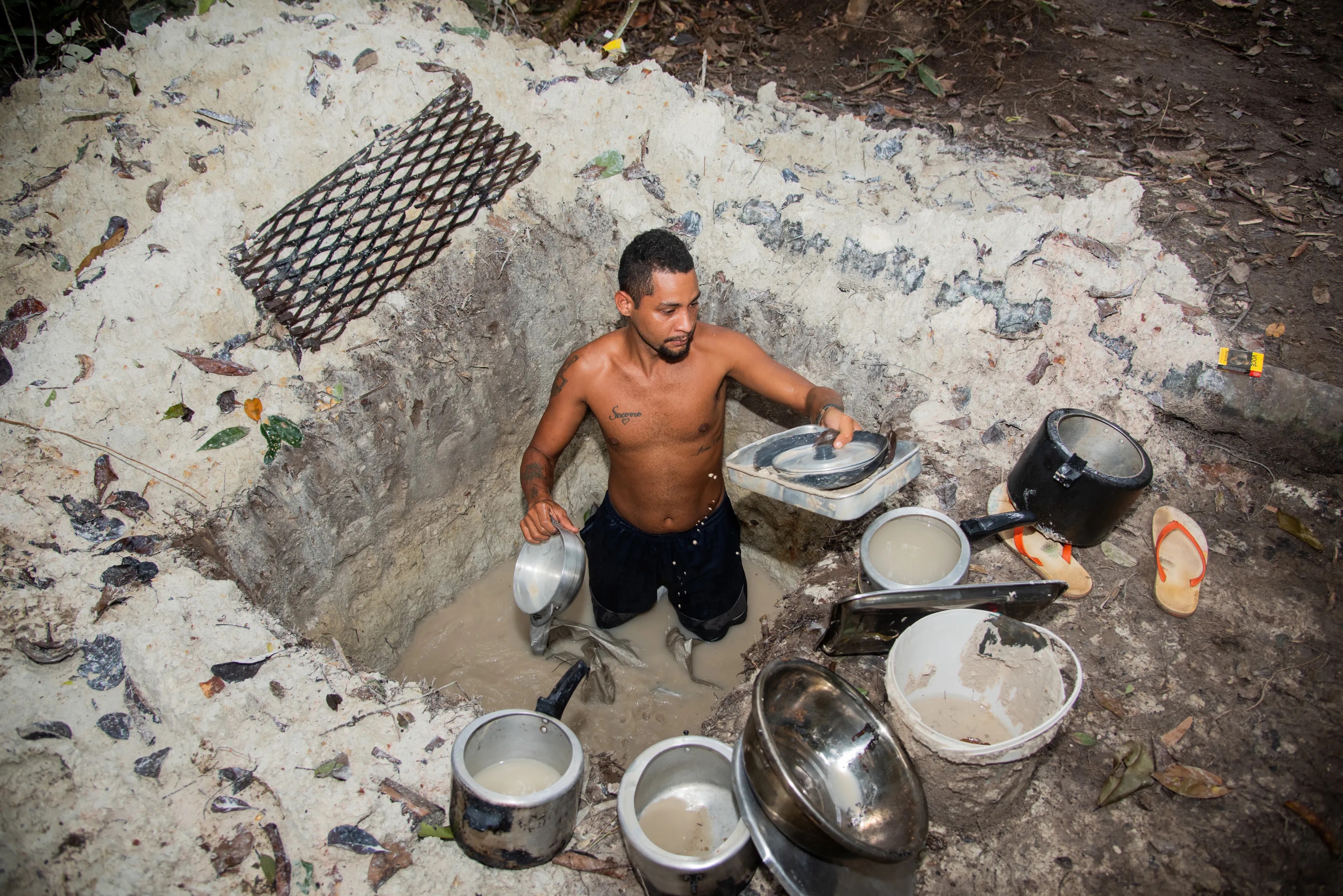 A man stands in a square-shaped pit, surrounded by gray earth and flooded with water. There are pots and pans on the edge of the pit, which is as deep as his elbows. The surrounding dirt is a darker gray-brown.