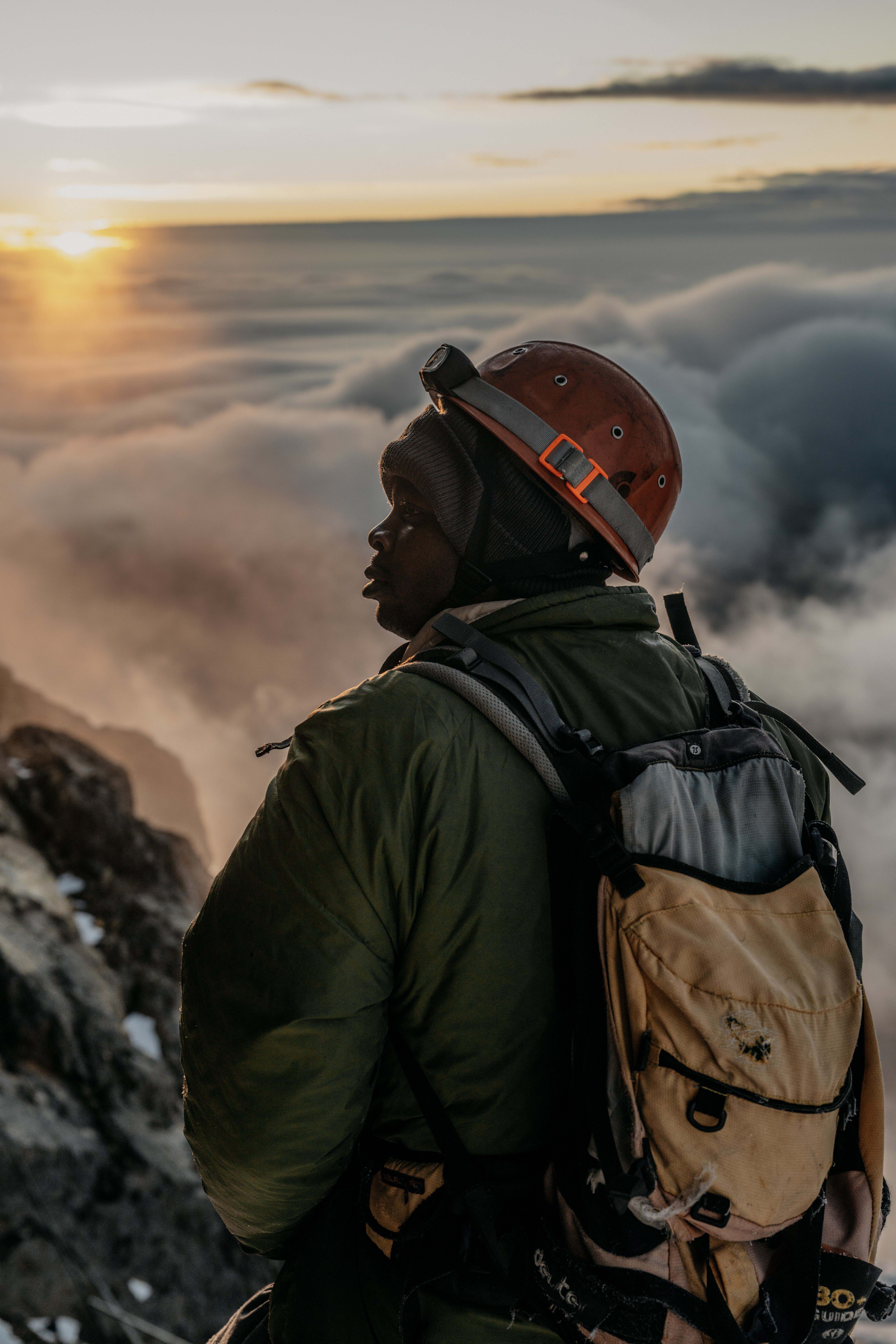 A mountain guide in full protective gear looks to the side as the sun meets the horizon above the clouds.