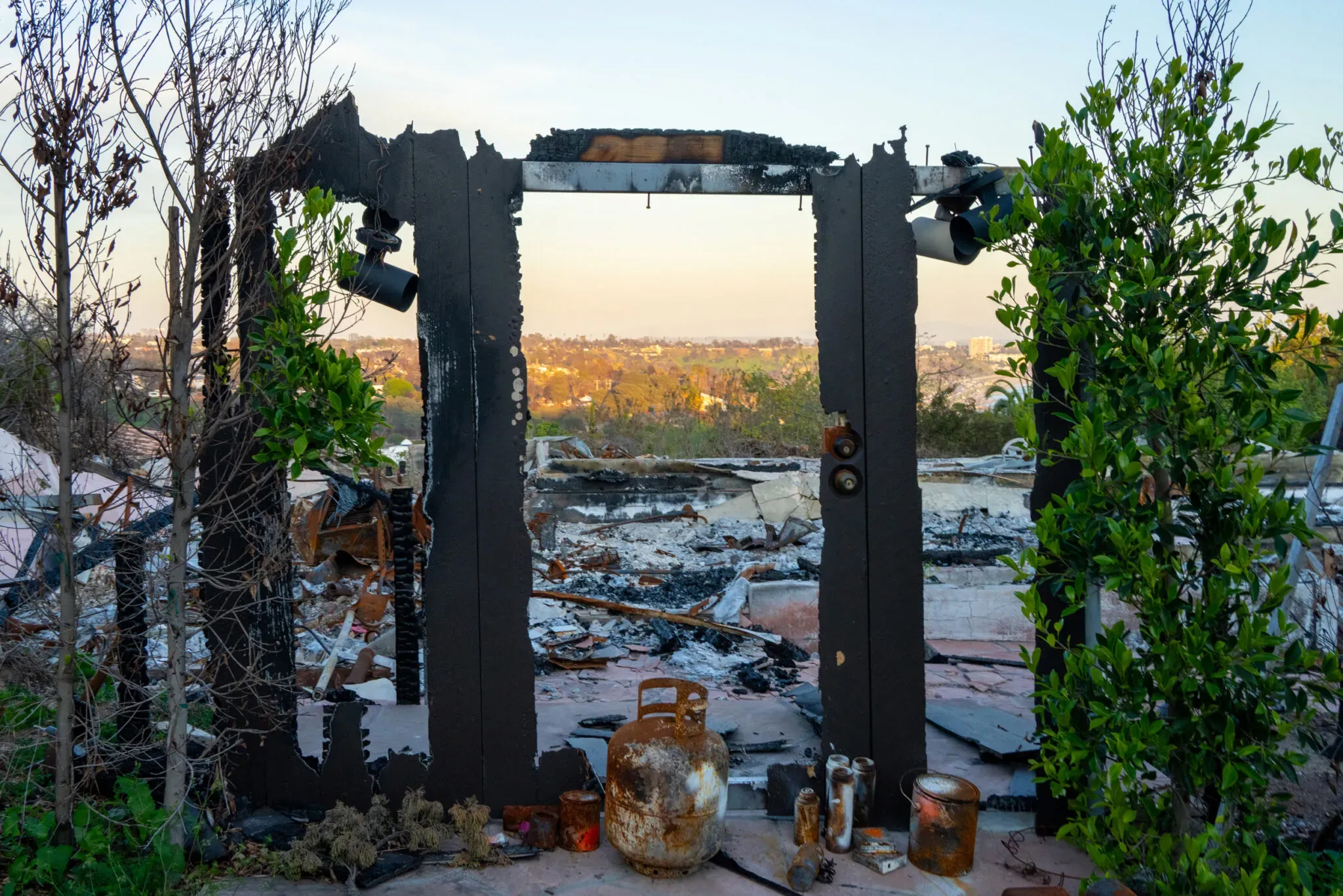 In the foreground, a charred door frame is flanked by green bushes. Behind the frame are burned ruins of a household.