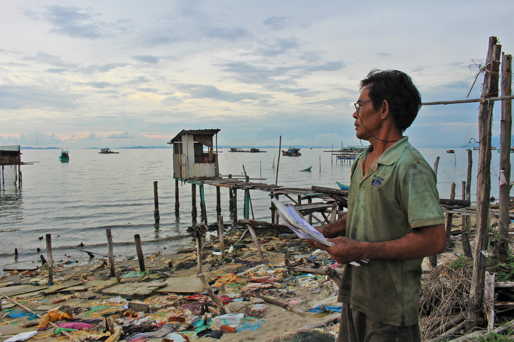 A man holds paperwork as he looks over the water. Debris from demolished homes are littered on the shore.