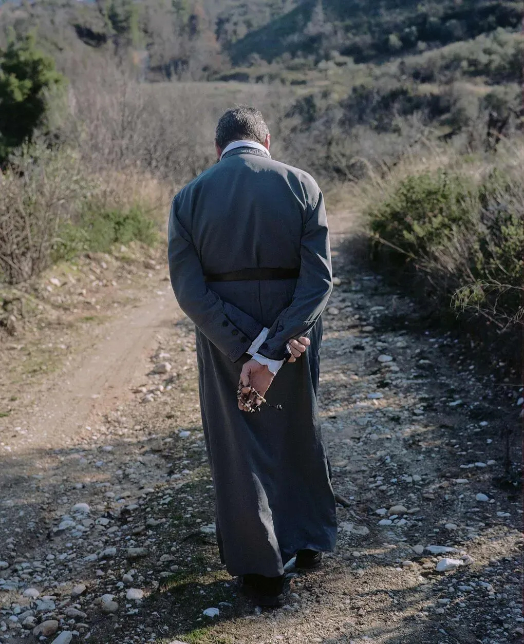 An older man walks down a dirt road with his hands behind his back, seen from behind. The road is surrounded by olive groves.