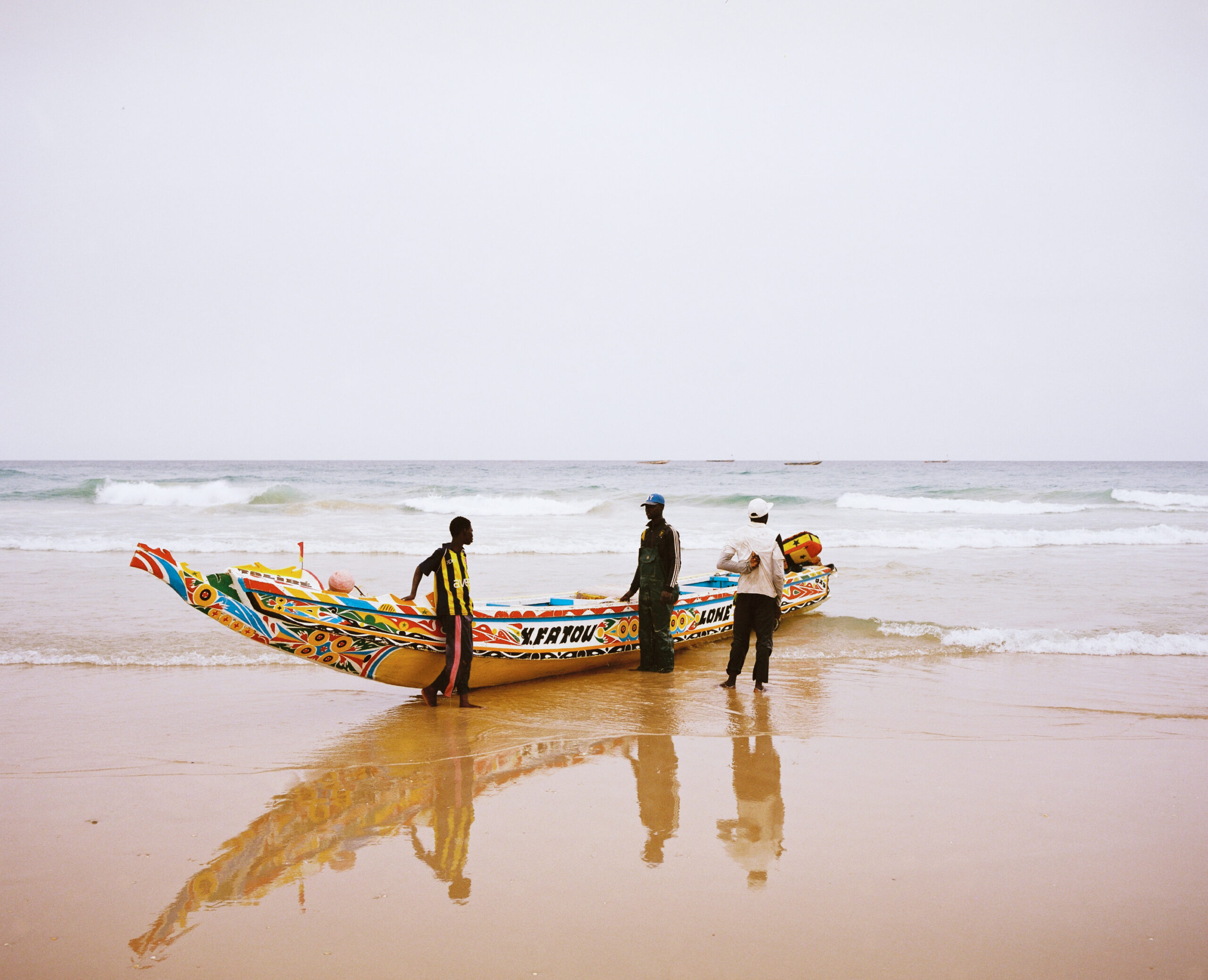 Three fishermen stand by their colorful decorated boat on a beach, their shadows reflected in the wet sand. In the background are ocean waves and an overcast sky.