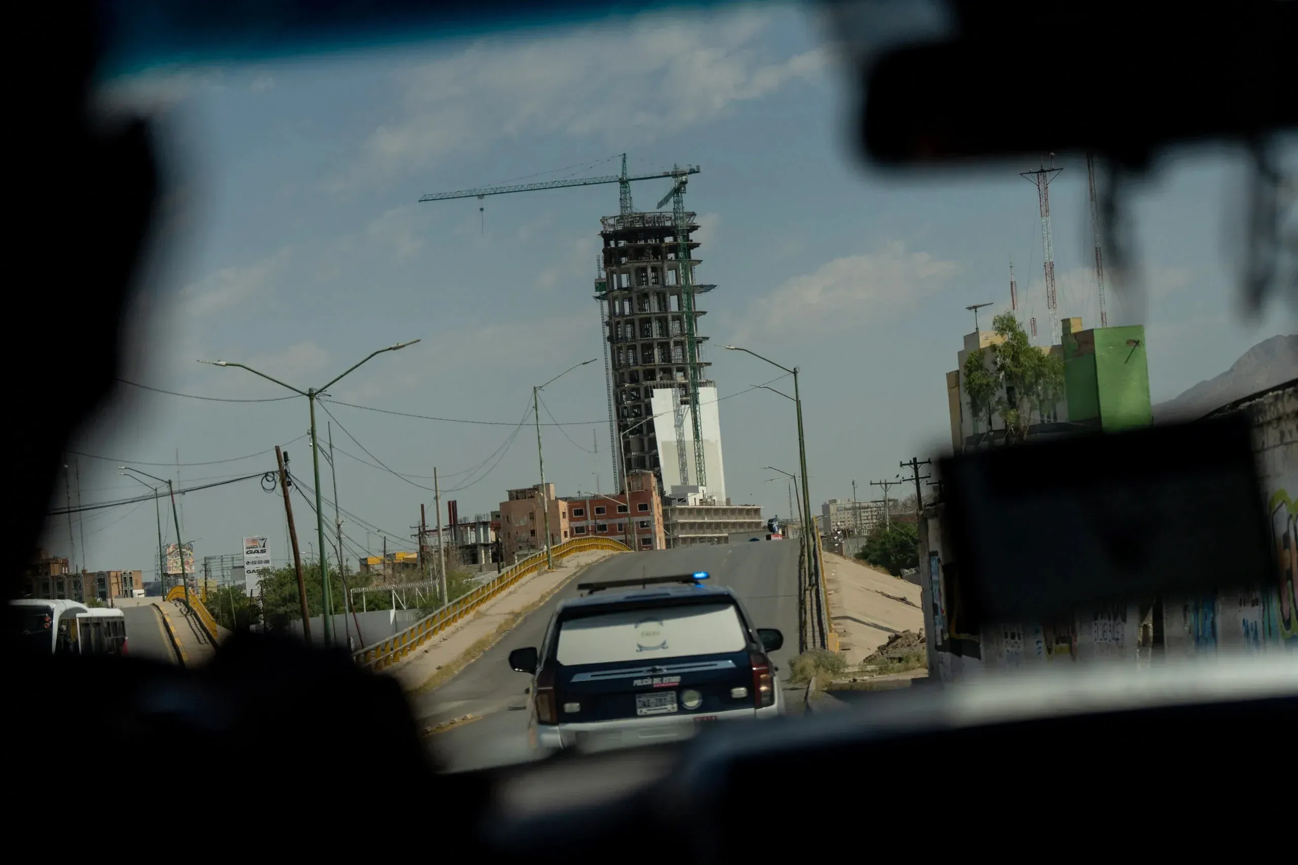 View through a car windshield of a police SUV with flashing lights driving on an overpass toward a tall, unfinished high-rise under construction, topped with a crane