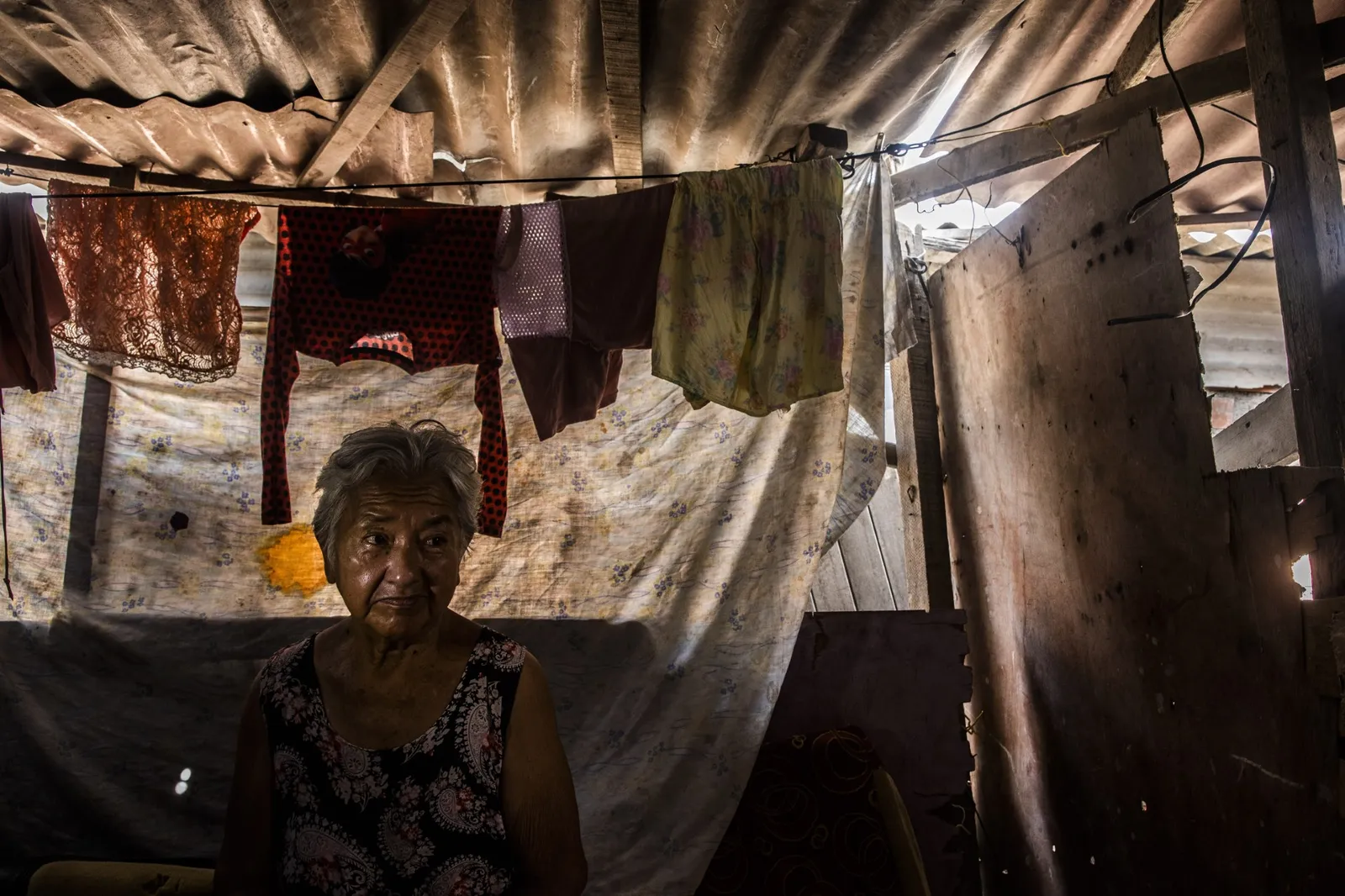 In a darkened area, an older woman sits in a structure made of cardboard and corrugated iron.