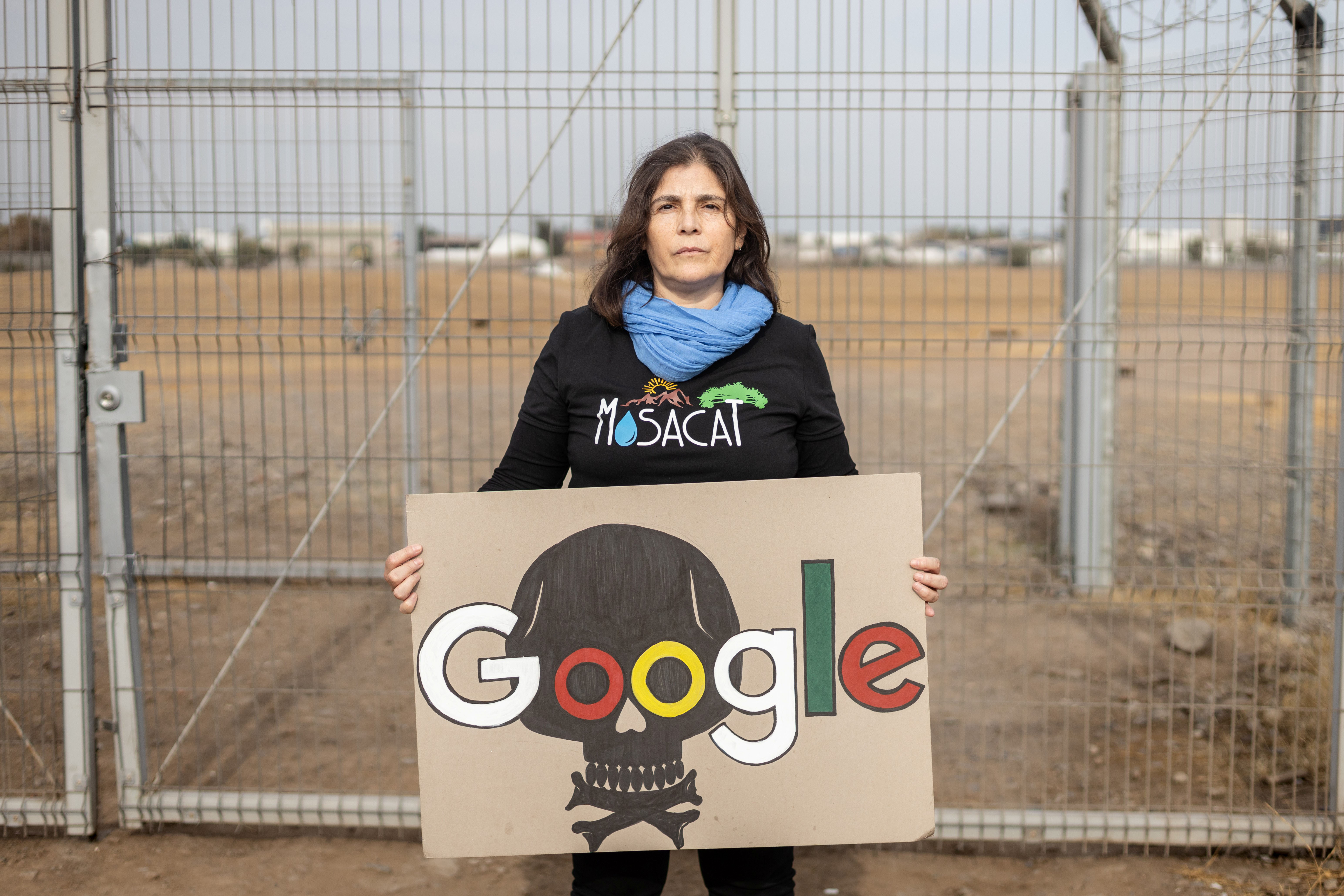 A woman wearing a dark shirt reading “Mosacat” and holding a handmade “Google” sign stands in front of a fence on a lot, with white buildings in the background.