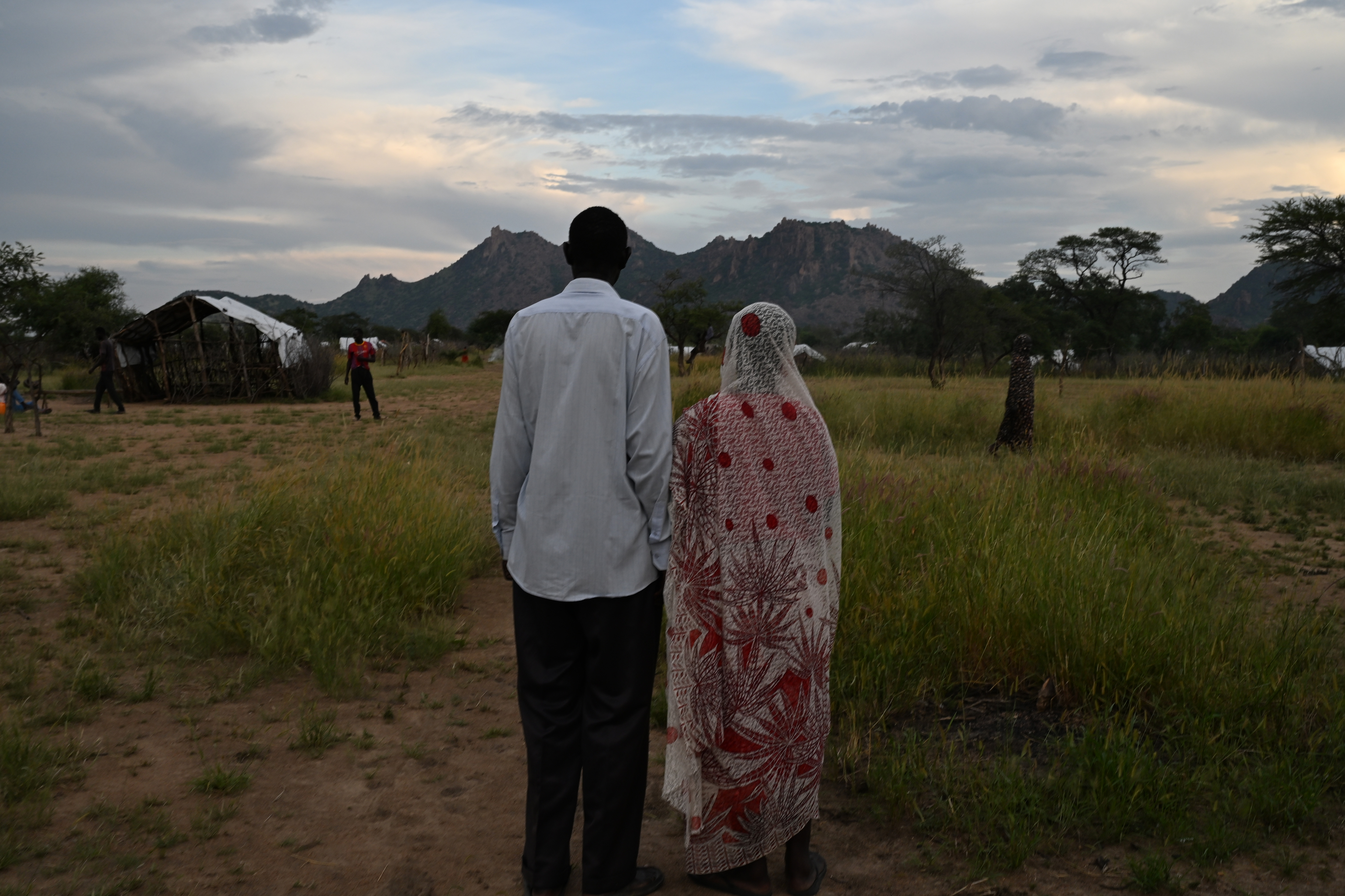 A couple stands on a grassy plain, their backs facing the camera. They are looking out at a mountain ridge in the distance. On the left, the man is wearing dark pants and a light shirt. On the right, the woman is wearing a white headscarf and shawl with red stitching.