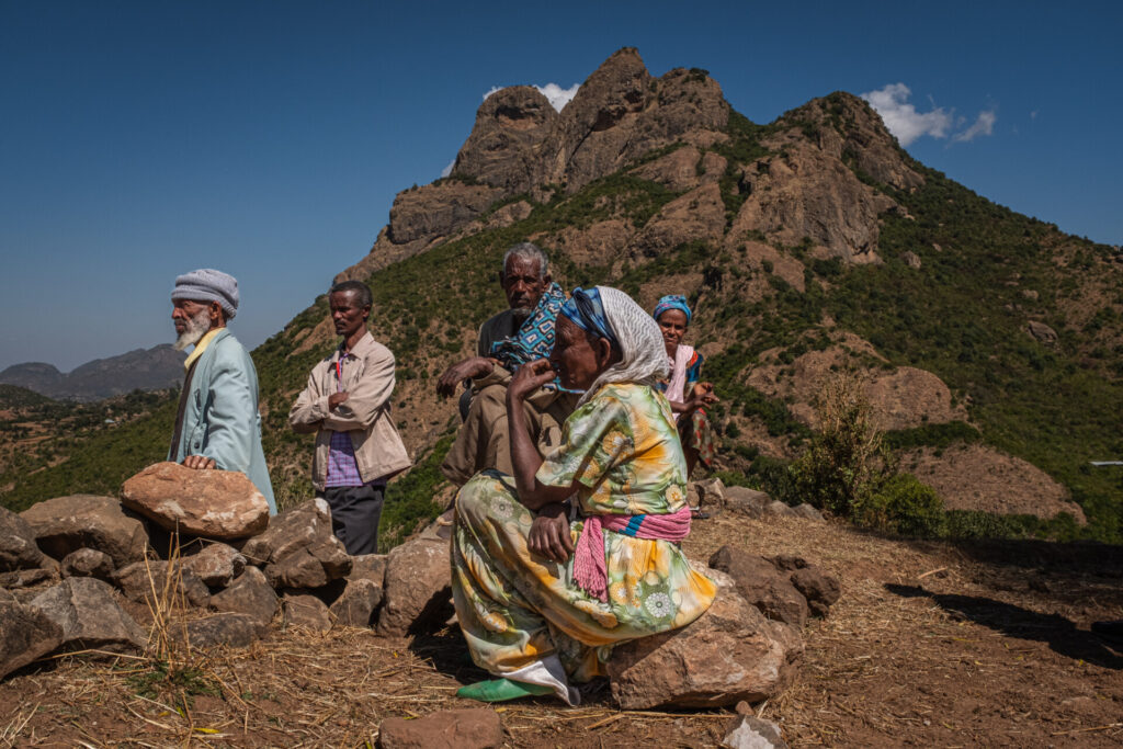 Five people sit and stand on a rock face, facing to the left. The sky is blue with few clouds. They are wearing colorful traditional clothing.