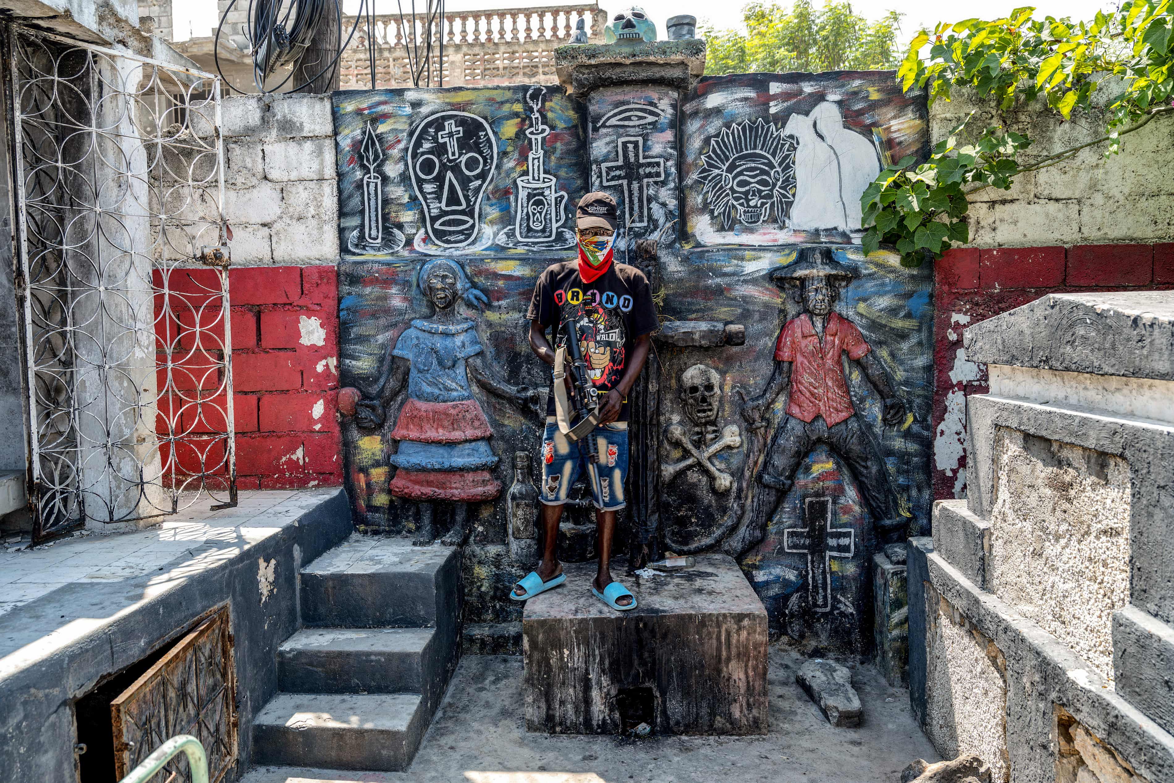 A man wearing dark clothing, a hat, and holding a gun stands in front of a wall filled with graffiti, almost blending into the graffiti background.
