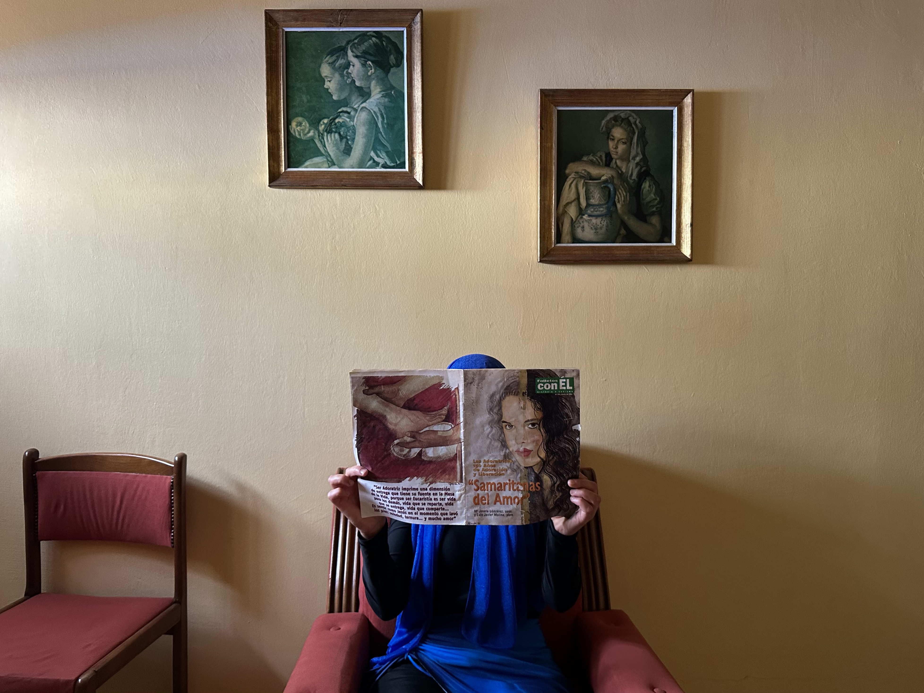 A woman in a bright blue hijab poses behind a magazine titled Samaritans of Love, hiding her face to conceal her identity. Behind her, framed paintings of women decorate the wall.