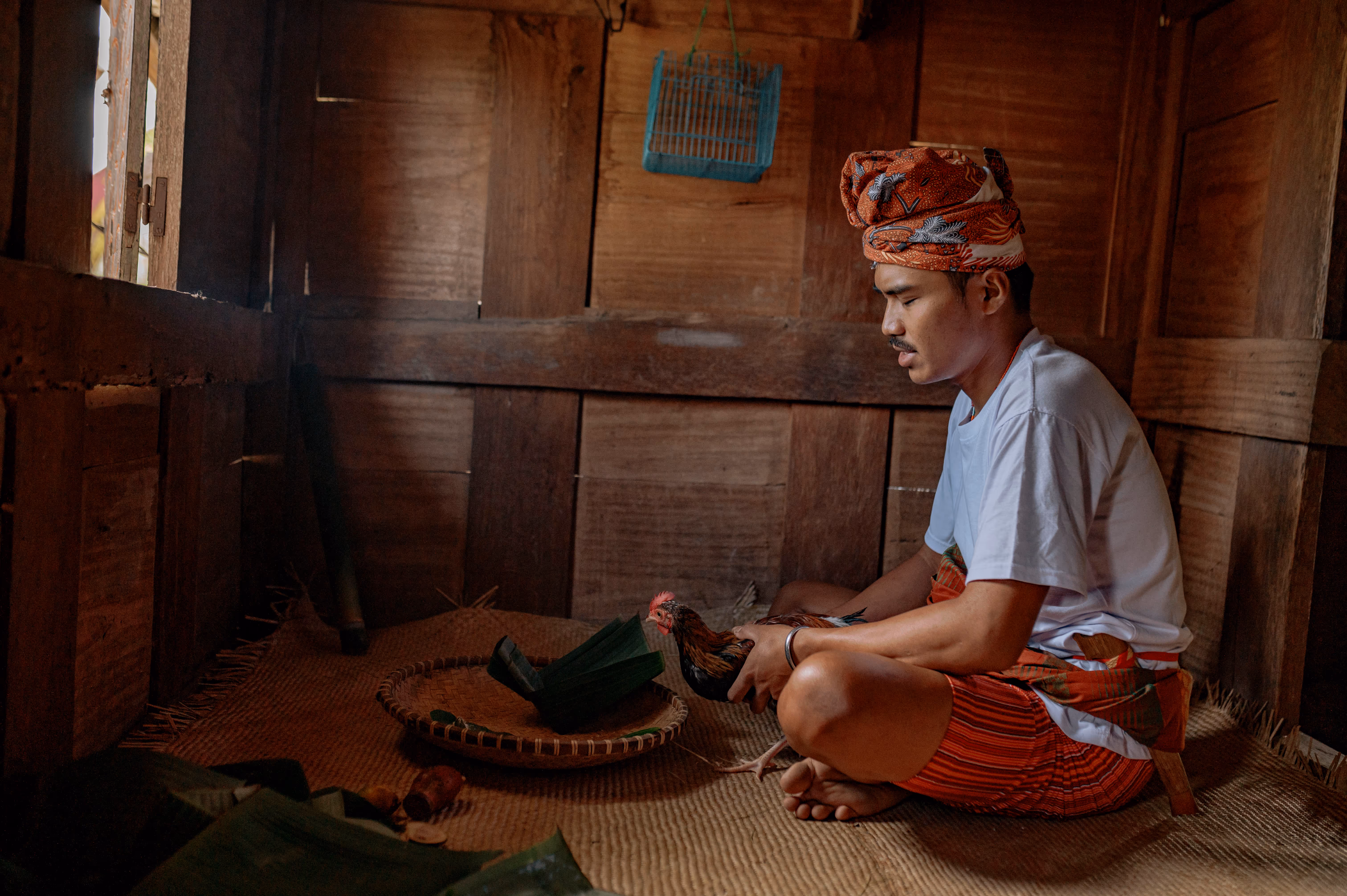 A young man sits with his legs folded in front of him and prays while holding a chicken.