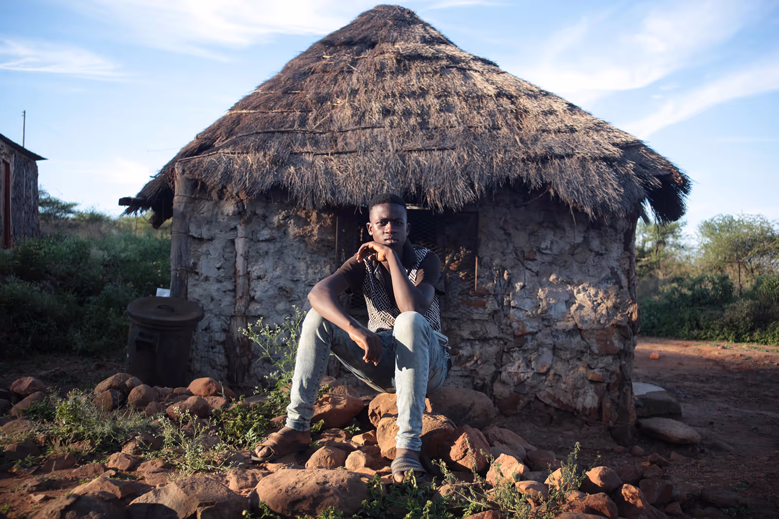 A teenager sits on a rock in front of a hut with a pensive look on his face. 