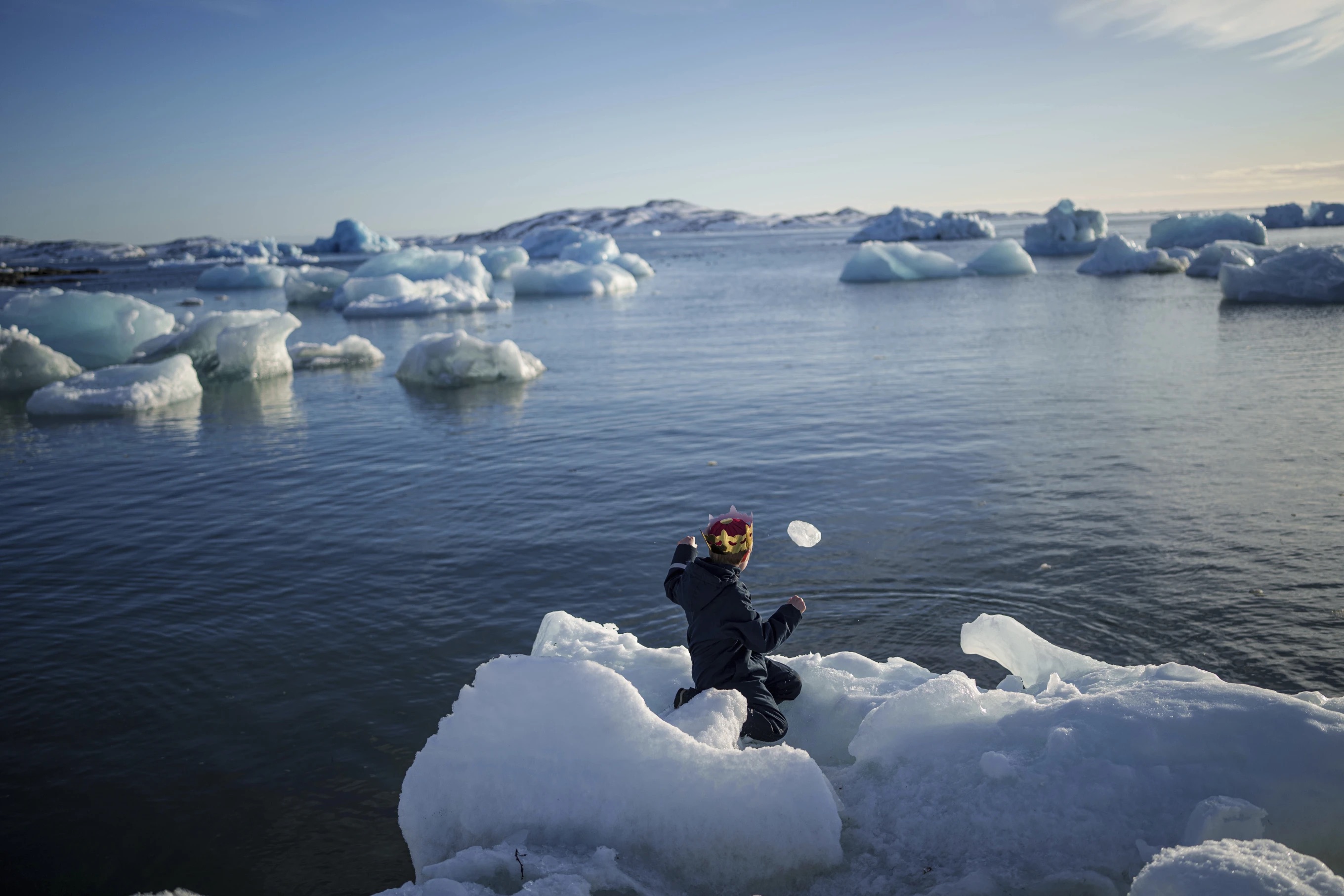 A child sits on a floating piece of ice as he throws a smaller piece of ice into the water. 