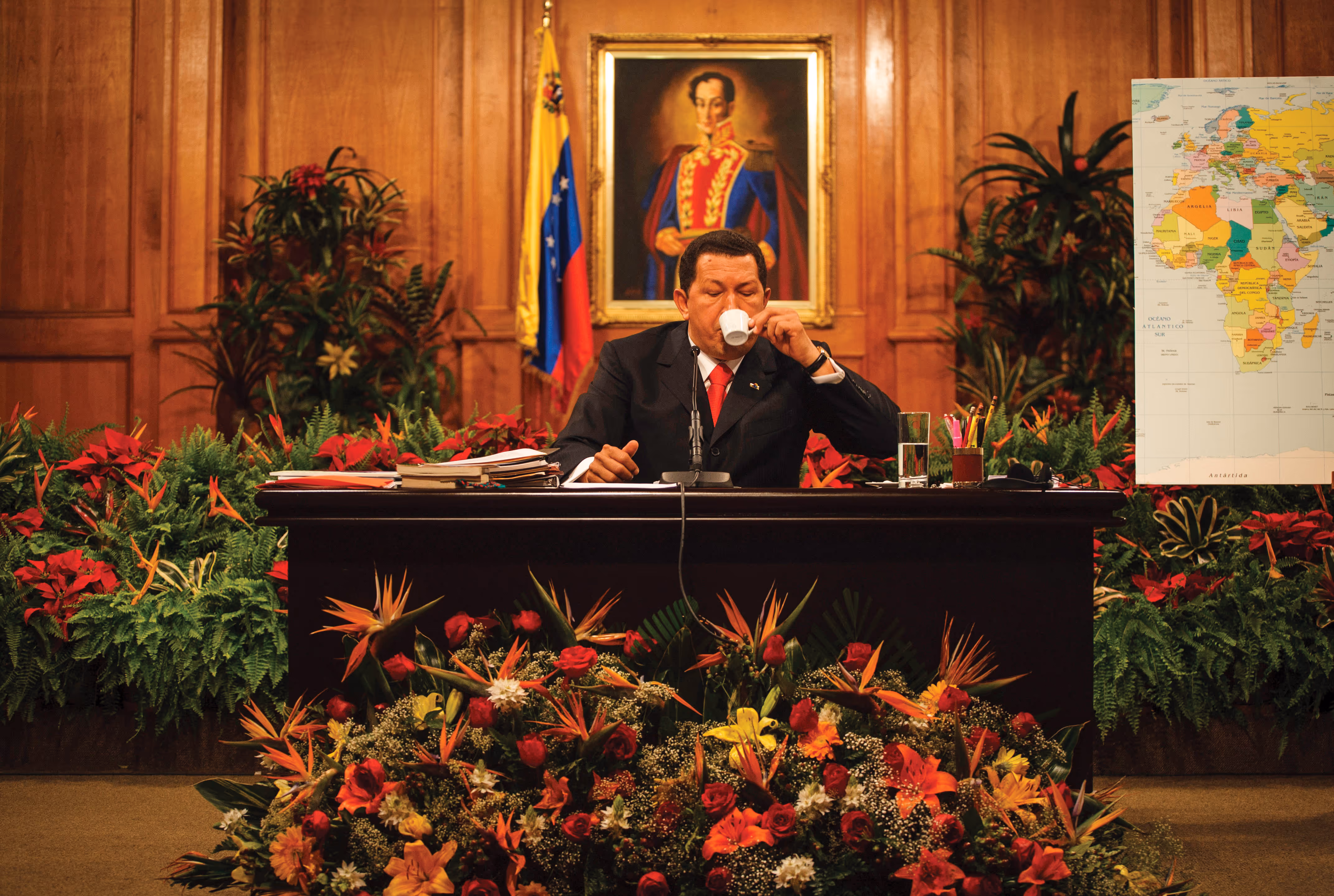 A man sits behind a desk surrounded with flowers and sipping an espresso cup