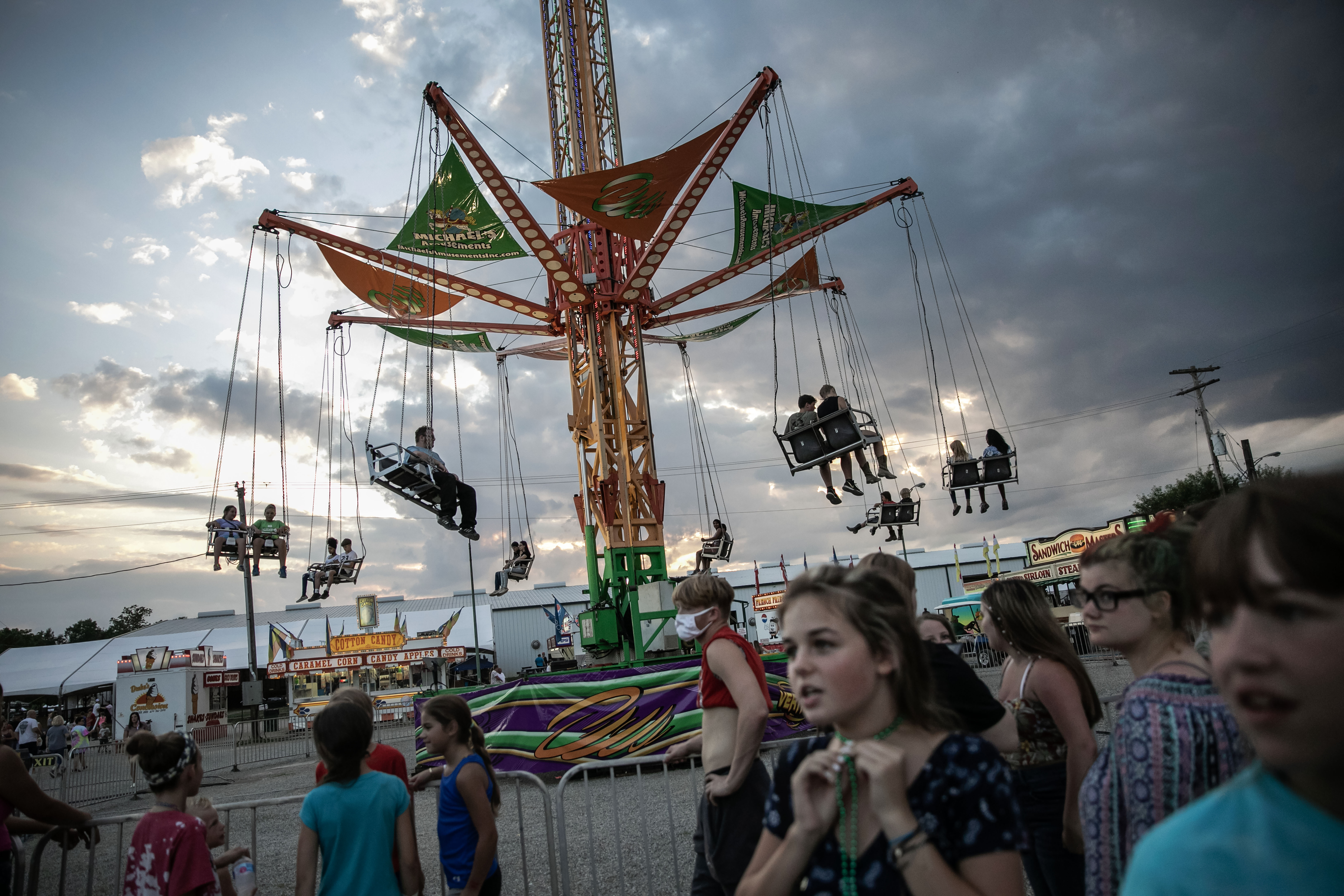 An amusement park ride runs in the background; young people without masks walk in the foreground