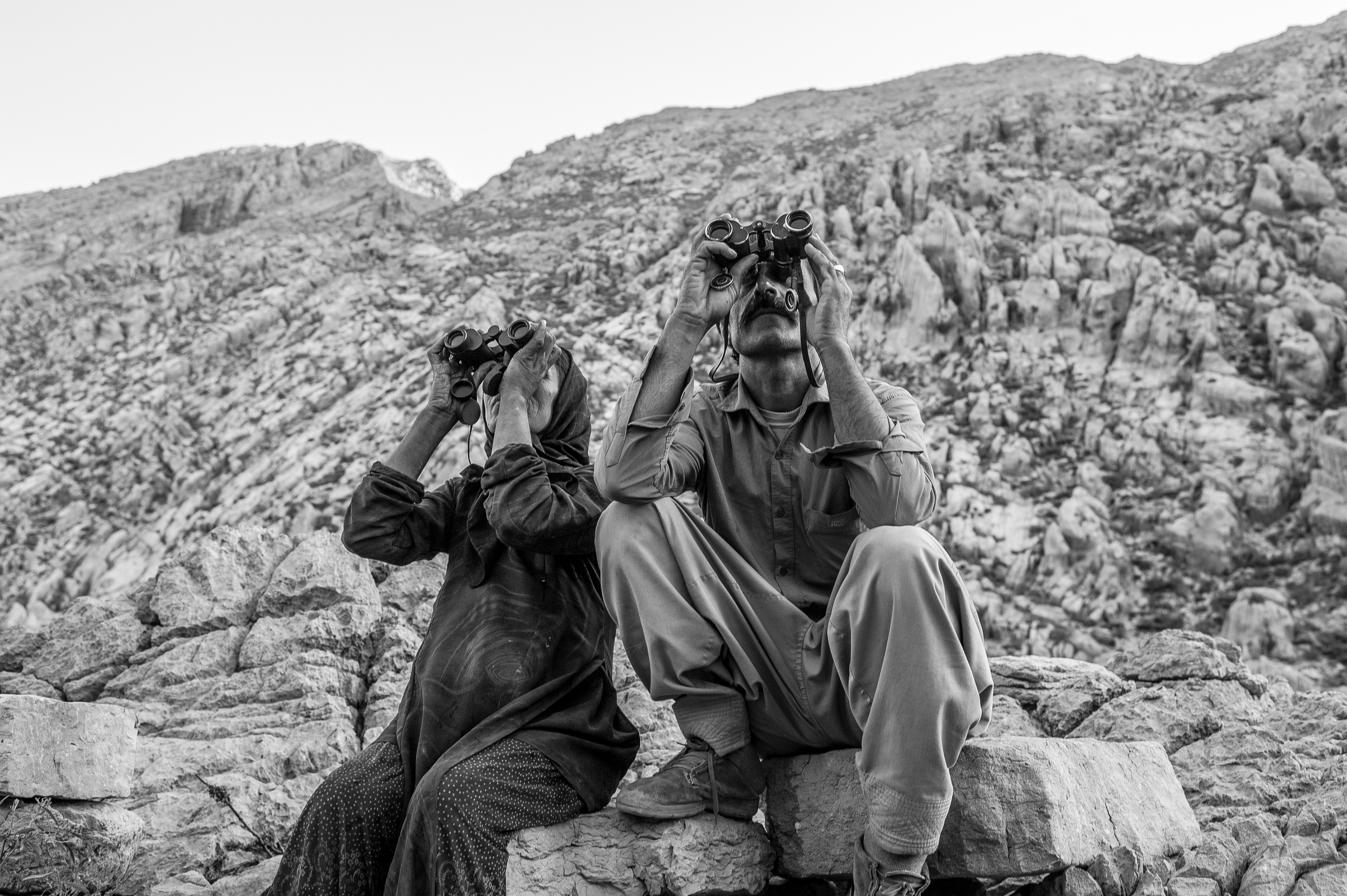 Two people sit on a rock with binoculars trained at the sky
