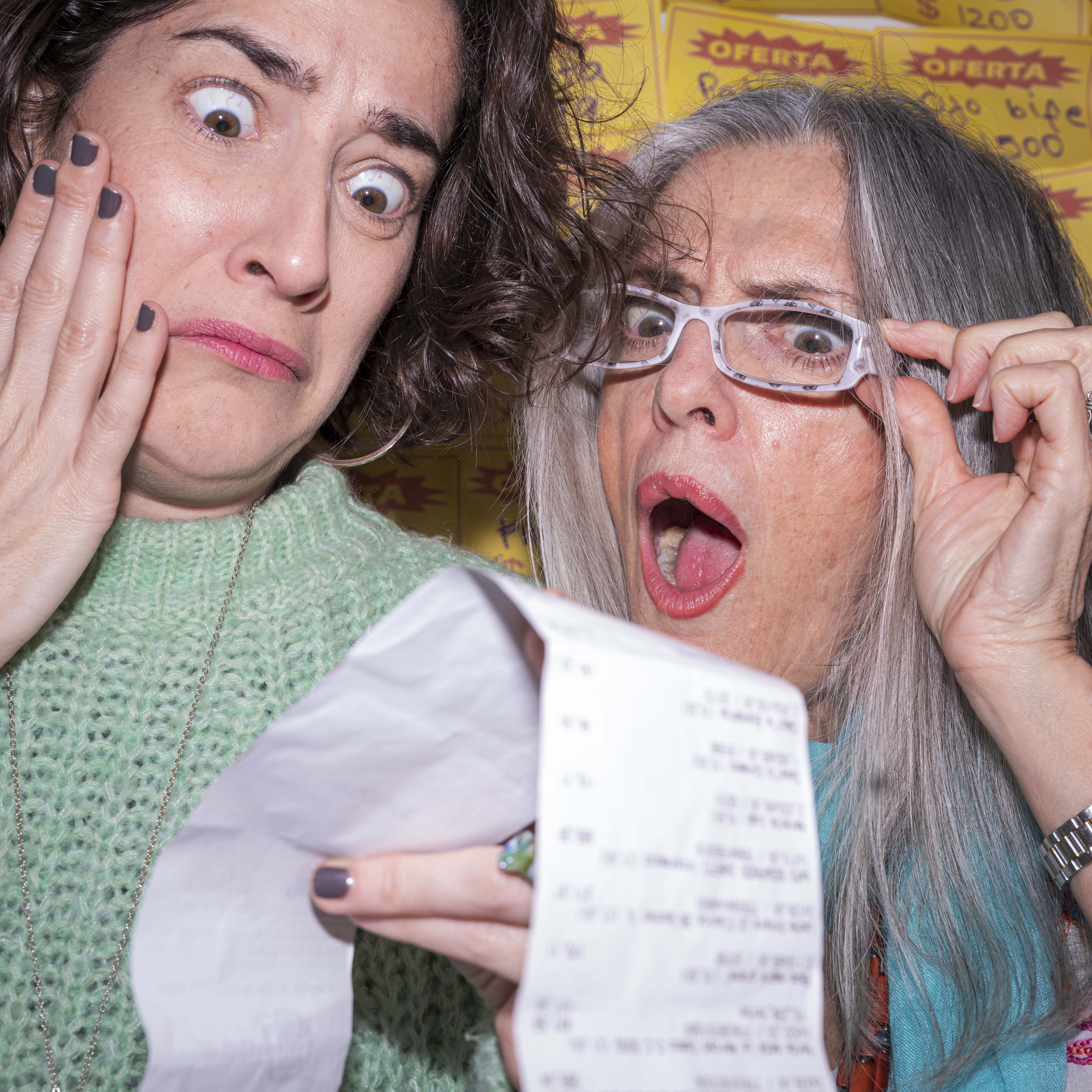 Two women look at a receipt making comical expressions