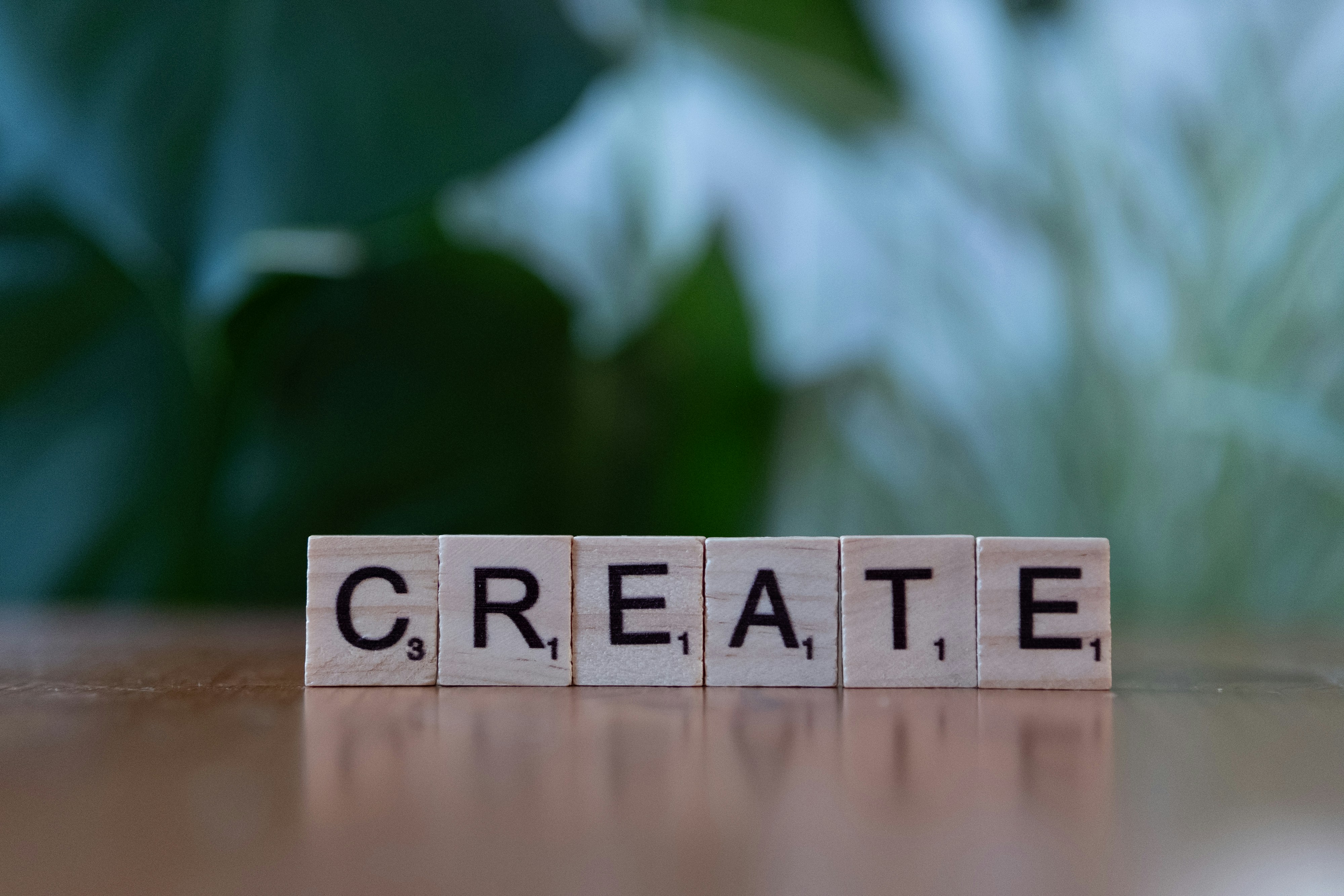 Wooden Scrabble tiles arranged to spell the word CREATE on a wooden surface with an out-of-focus green leafy background.