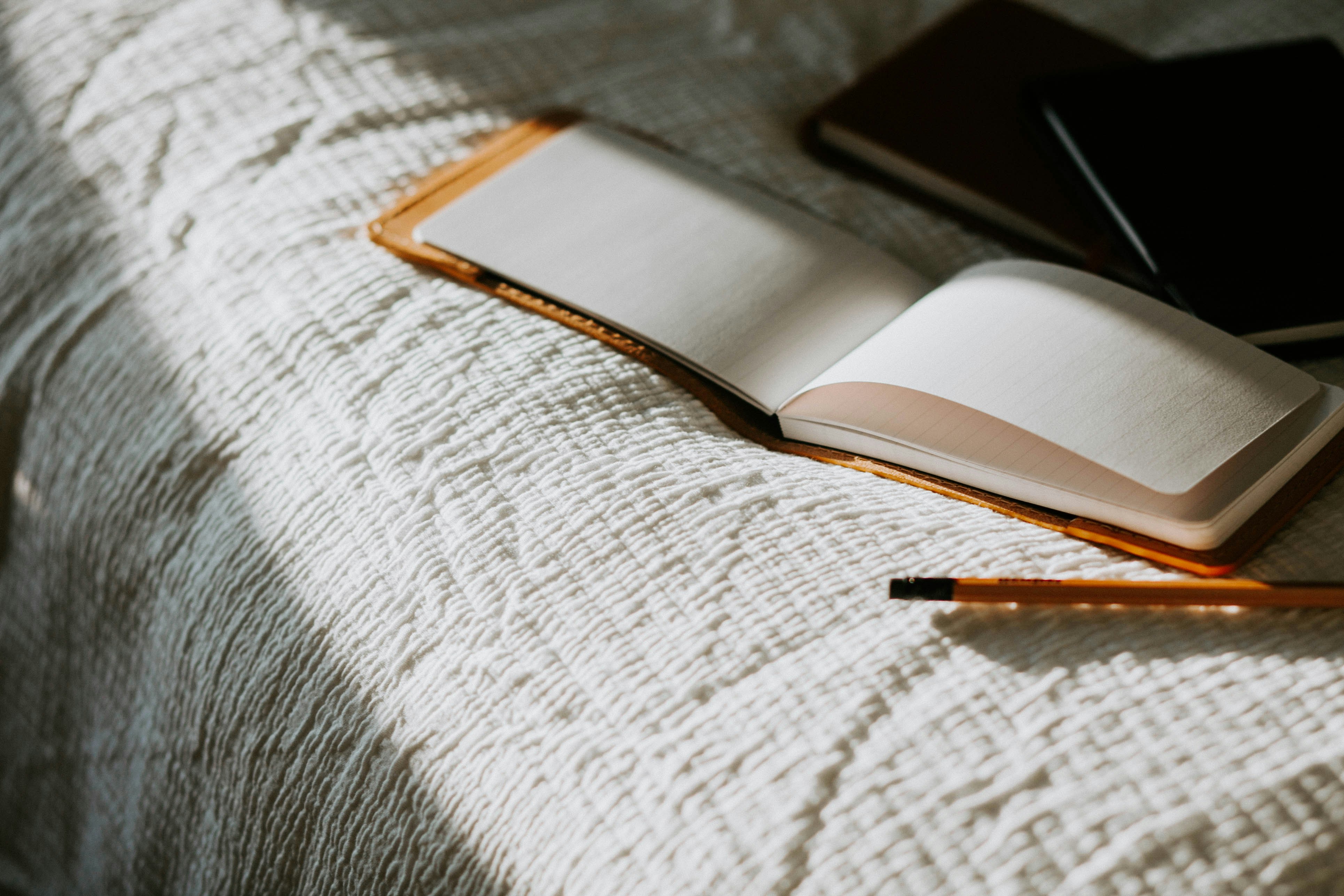 Open blank notebook with a brown cover on a textured white bedspread, accompanied by two closed black notebooks and a pencil lying beside it.
