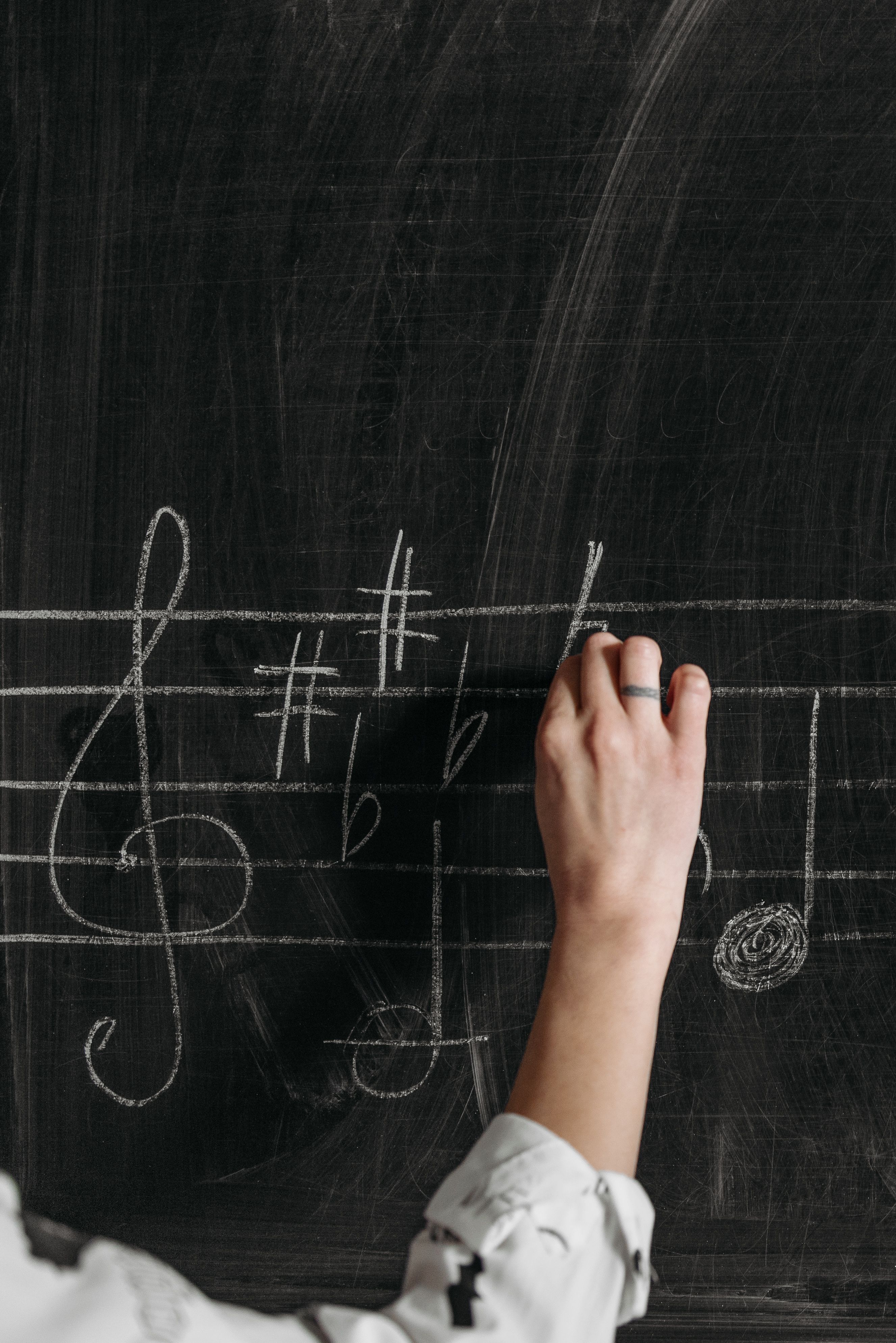 A person's hand writing musical notes and symbols on a blackboard with chalk, including a treble clef, sharps, flats, and a music note.