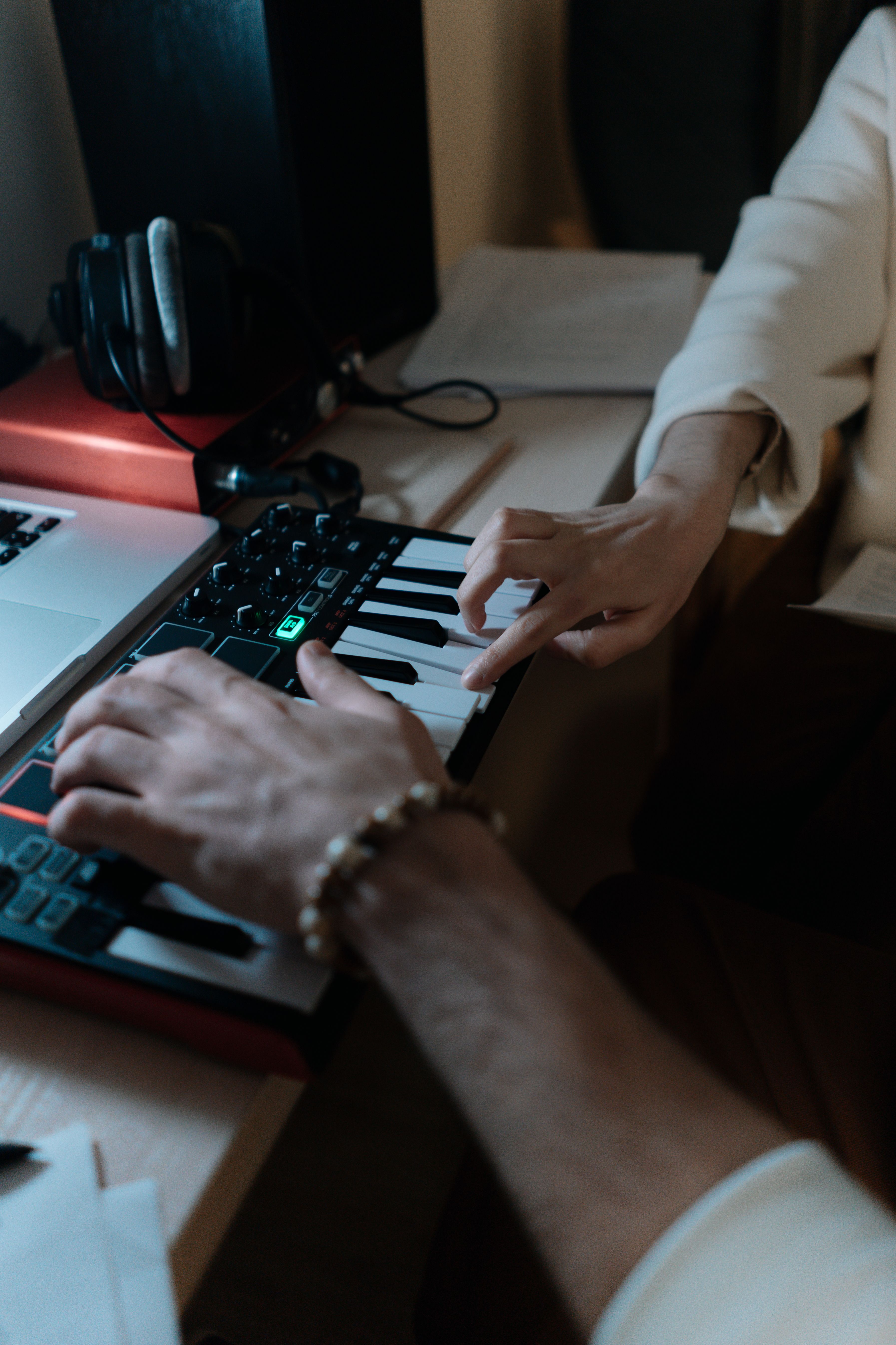 Two people using a small MIDI keyboard controller, one person's hands playing keys while the other points to a key, with a laptop and headphones nearby on the desk.