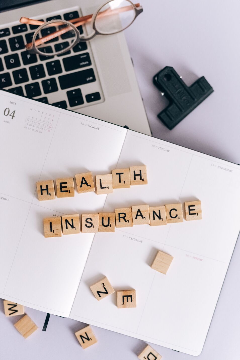Wooden Scrabble tiles spelling out 'HEALTH INSURANCE' placed on an open planner, with a pair of glasses resting on a laptop keyboard and a black binder clip nearby.