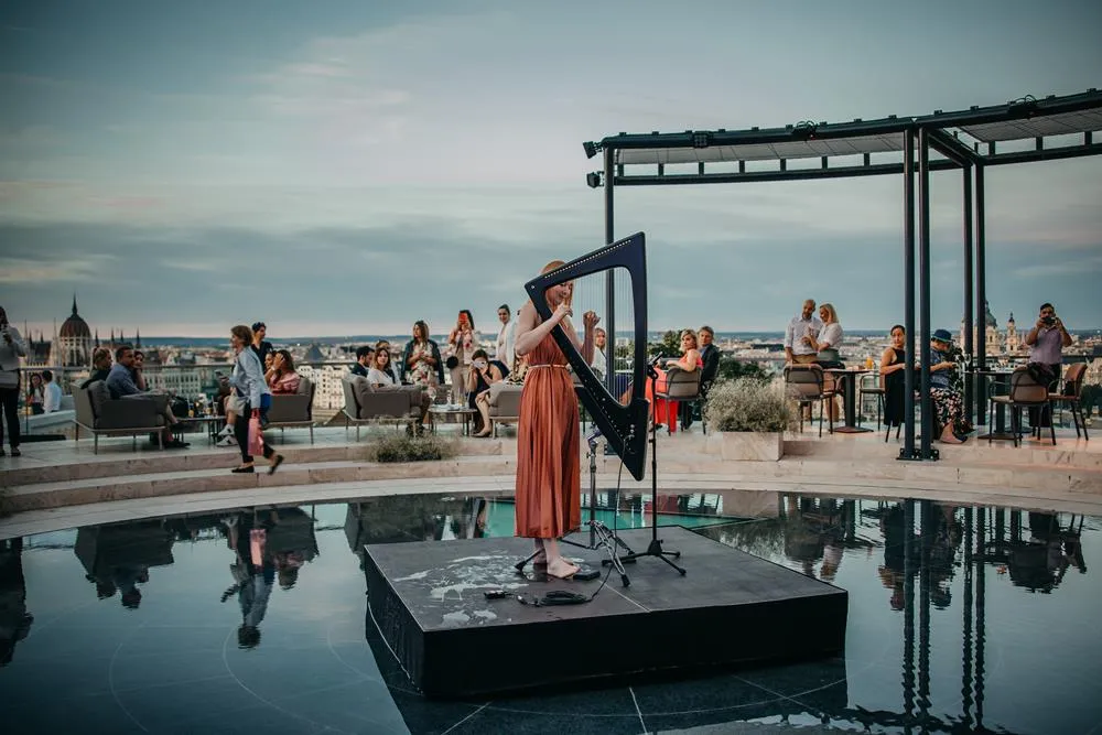 Álomszerű pillanat Budapest felett a naplementében, ahol egy elegáns hölgy hárfán játszik az Aranybástya teraszának feszített víztükrén.