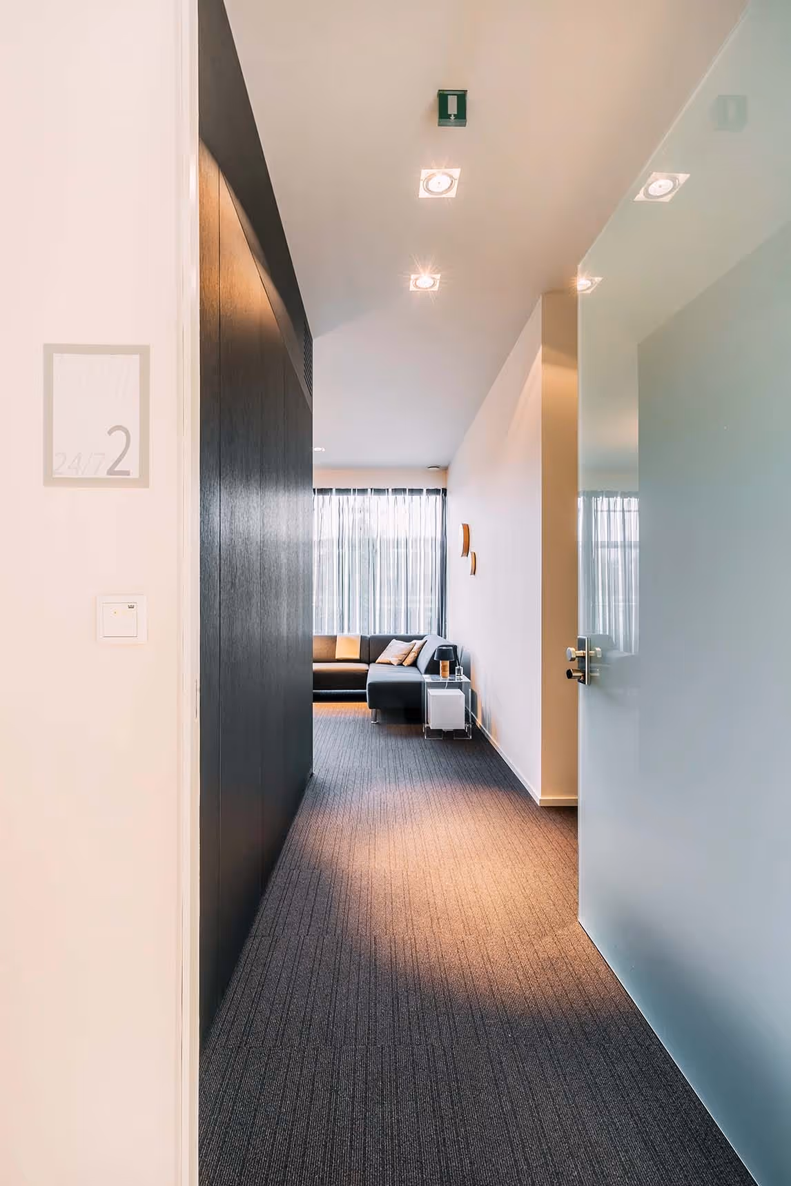 Doorkijk naar een modern ingerichte kamer met een grijze loungebank en zachte verlichting, zichtbaar vanuit een openstaande glazen deur en een gang met donker tapijt. Op de muur staat het kamernummer 2, met de aanduiding “24/7”.
