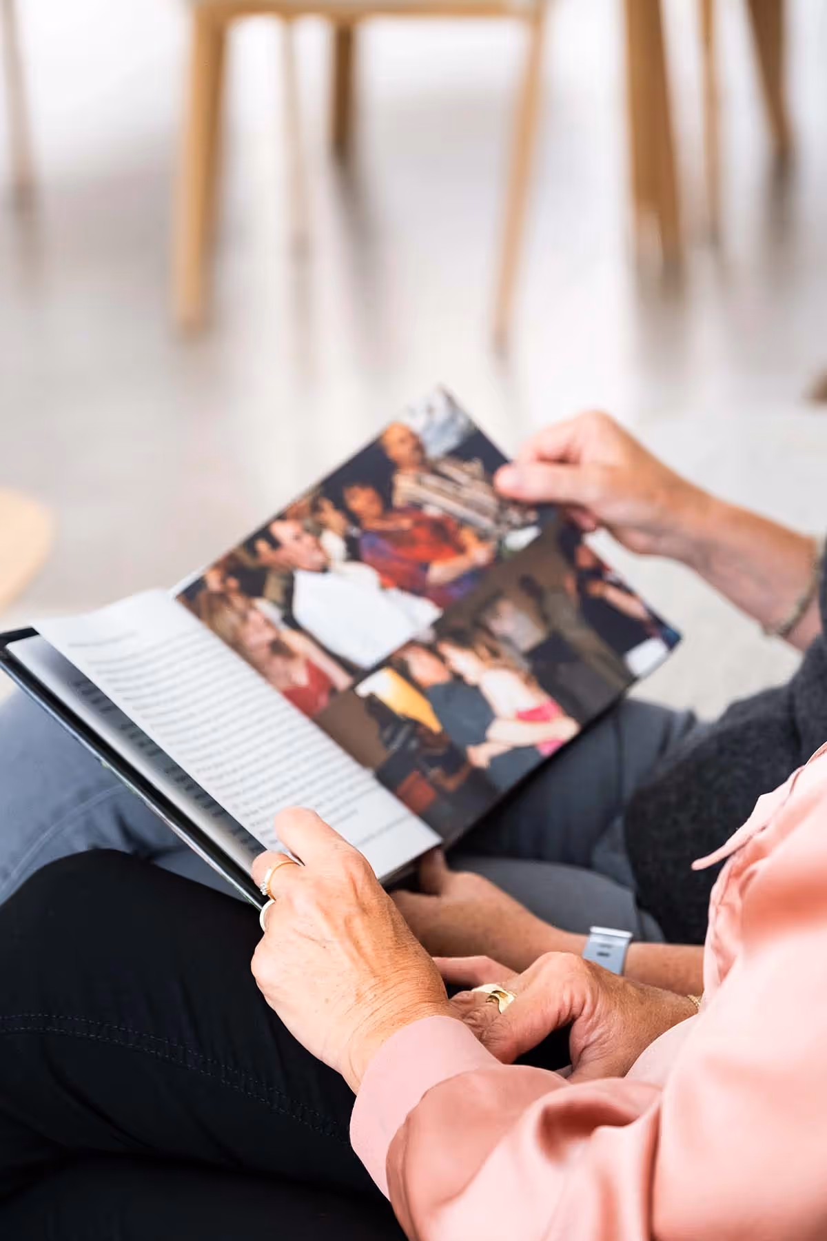 Twee oudere vrouwen zitten samen op een bank en bladeren door een fotoboek met herinneringen, waarin kleurrijke foto's van mensen op bijeenkomsten te zien zijn. Hun houding straalt rust en aandacht uit, in een lichte ruimte met houten stoelen op de achtergrond.
