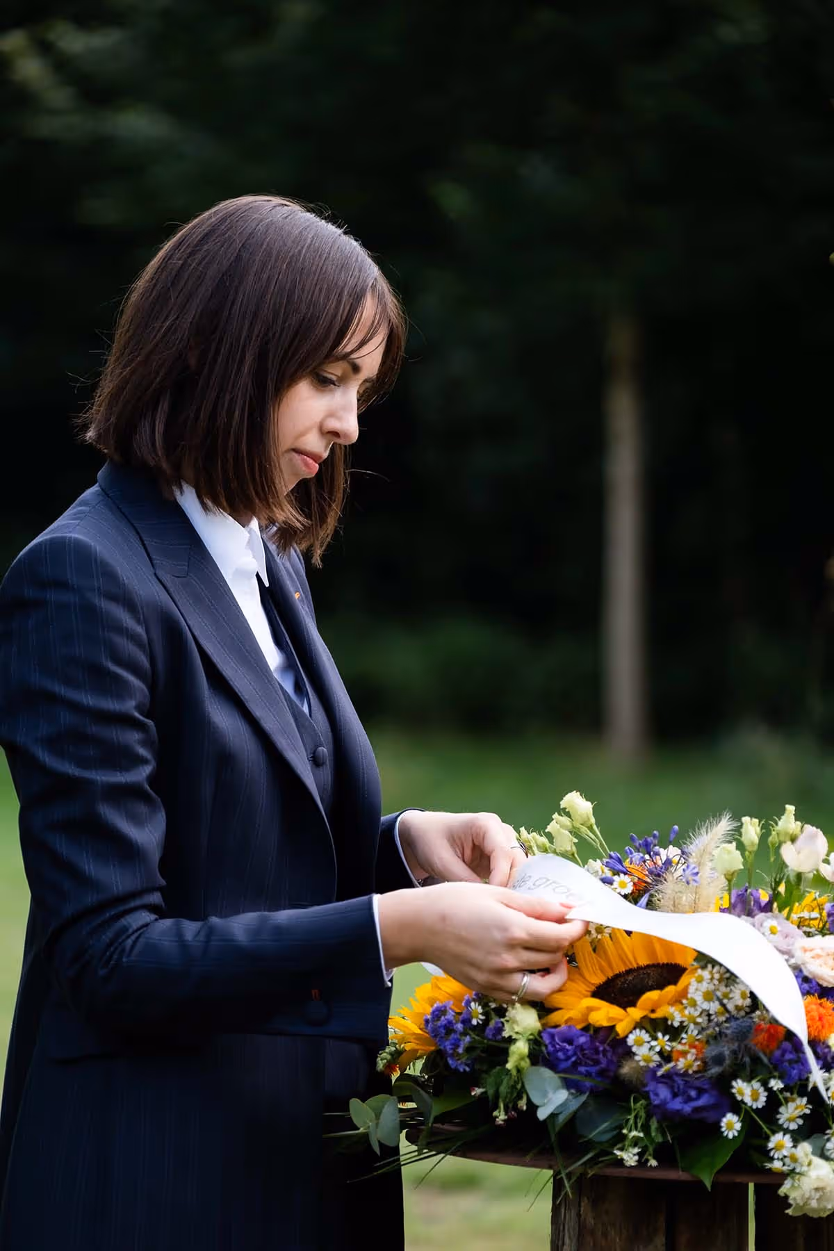 Een vrouw in een donkerblauw pak leest een lint op een kleurrijk rouwboeket met zonnebloemen, margrieten en paarse bloemen. Ze staat buiten op een grasveld, geconcentreerd en ingetogen.
