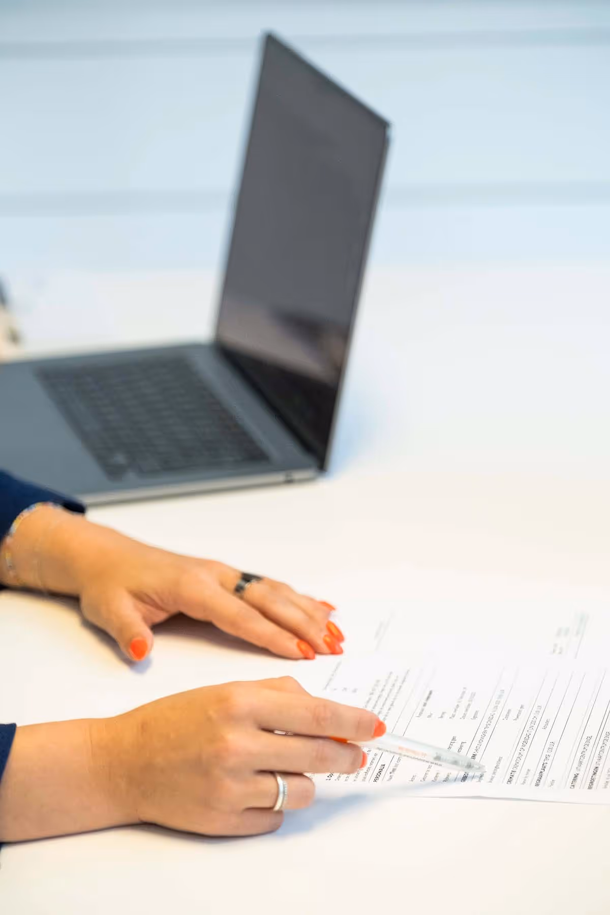 Close-up van twee handen met feloranje nagellak die een formulier op tafel invullen, met een opengeklapte laptop op de achtergrond. Eén hand wijst met een pen naar een tekstvak op het formulier.