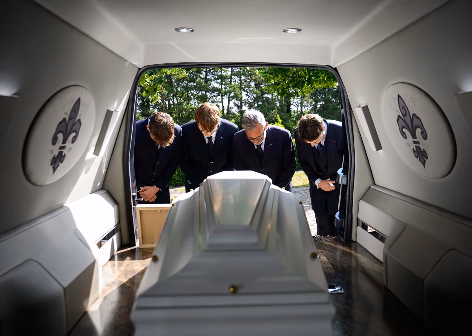 Zicht vanuit de achterkant van een lijkwagen op een witte kist, met vijf mannen in donkere pakken die eerbiedig buigen. Ze staan buiten in het zonlicht, omringd door groen, en tonen respect tijdens een uitvaartceremonie.