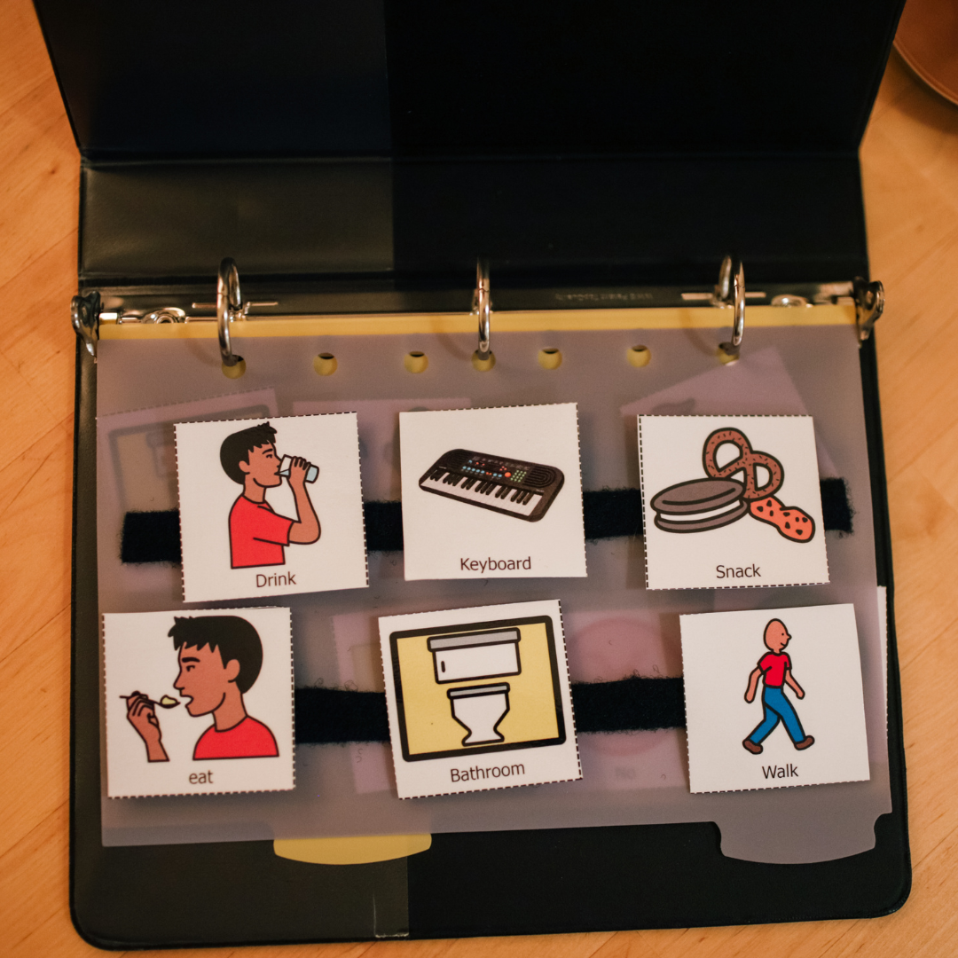Ring-bound AAC communication book showing picture symbols for drink, keyboard, snack, eating, bathroom, and walking