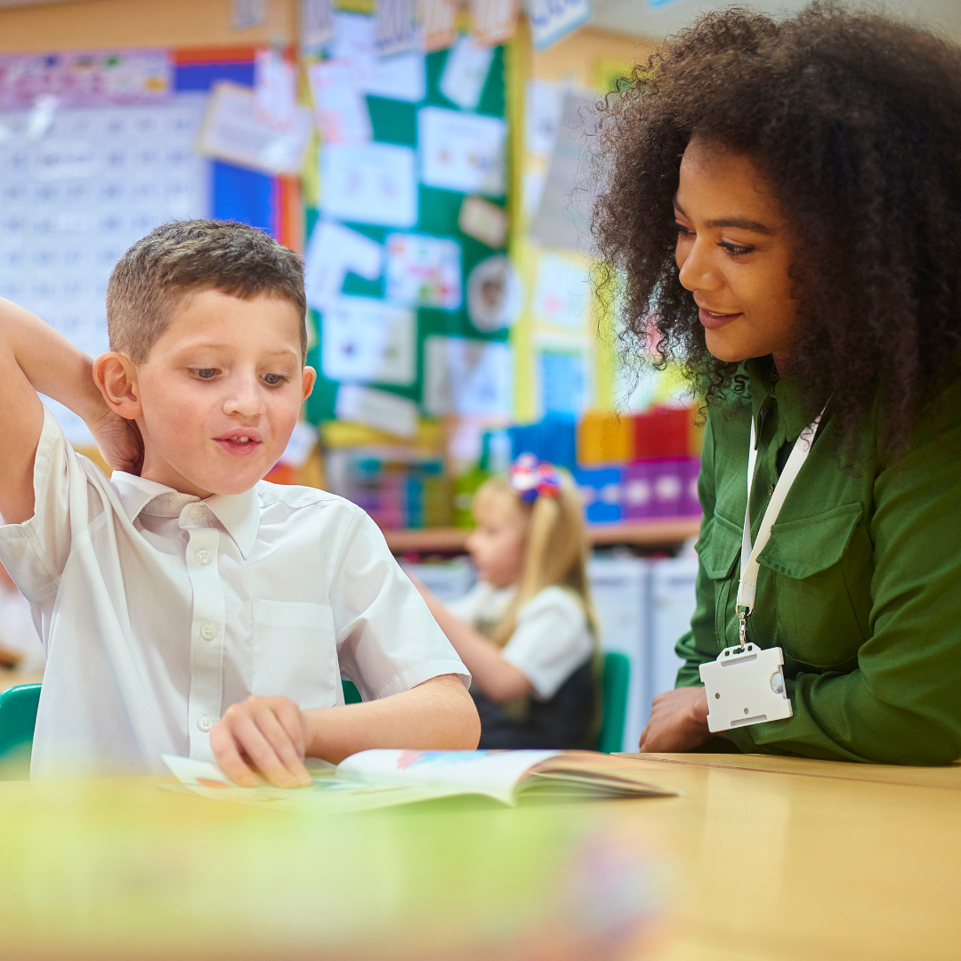 Teacher sitting beside a young student reading a book in a classroom
