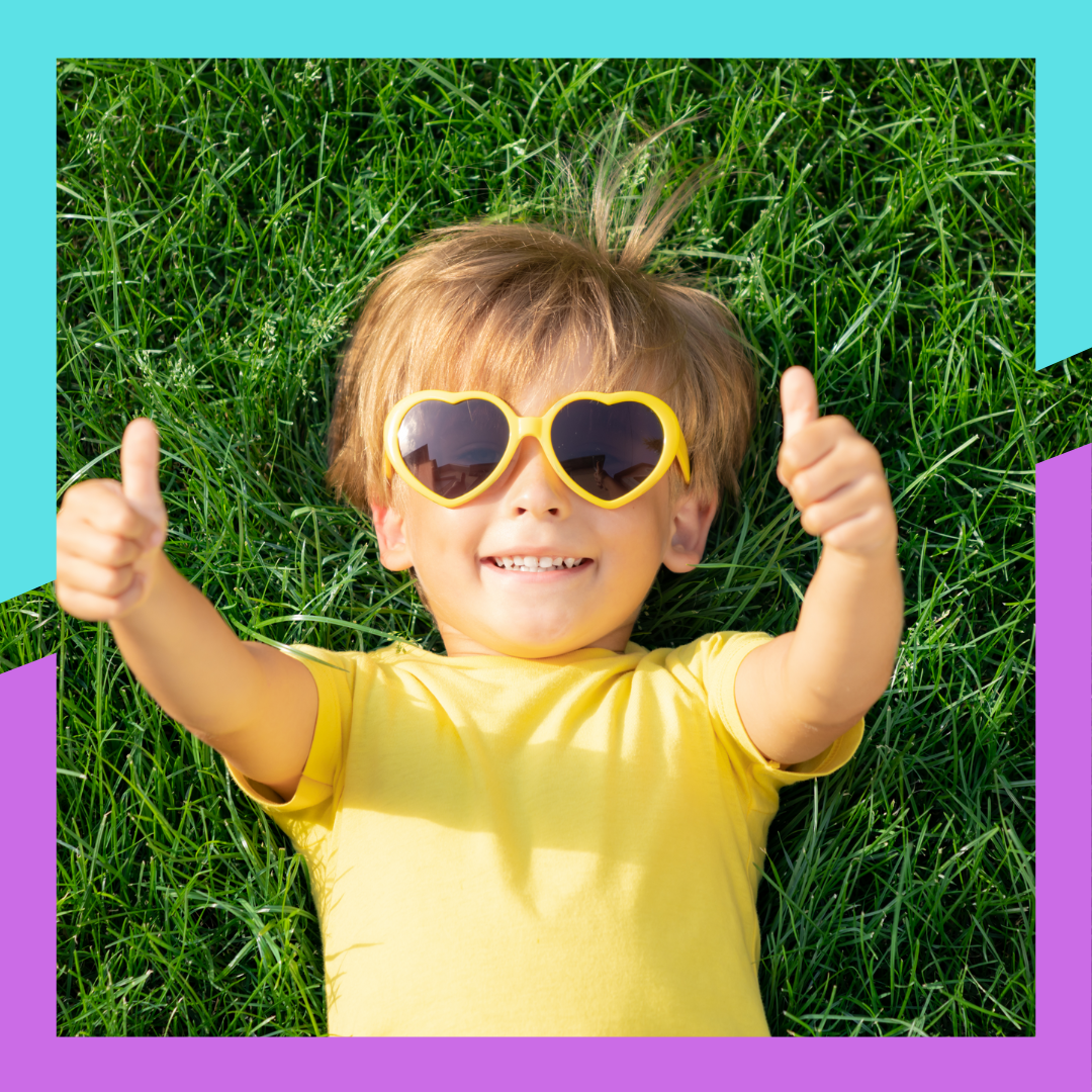 Smiling child in heart-shaped sunglasses giving two thumbs-up while lying on grass