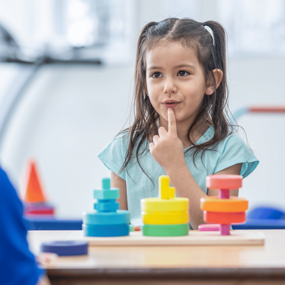 Young child touching her lips while playing with colorful stacking toys.