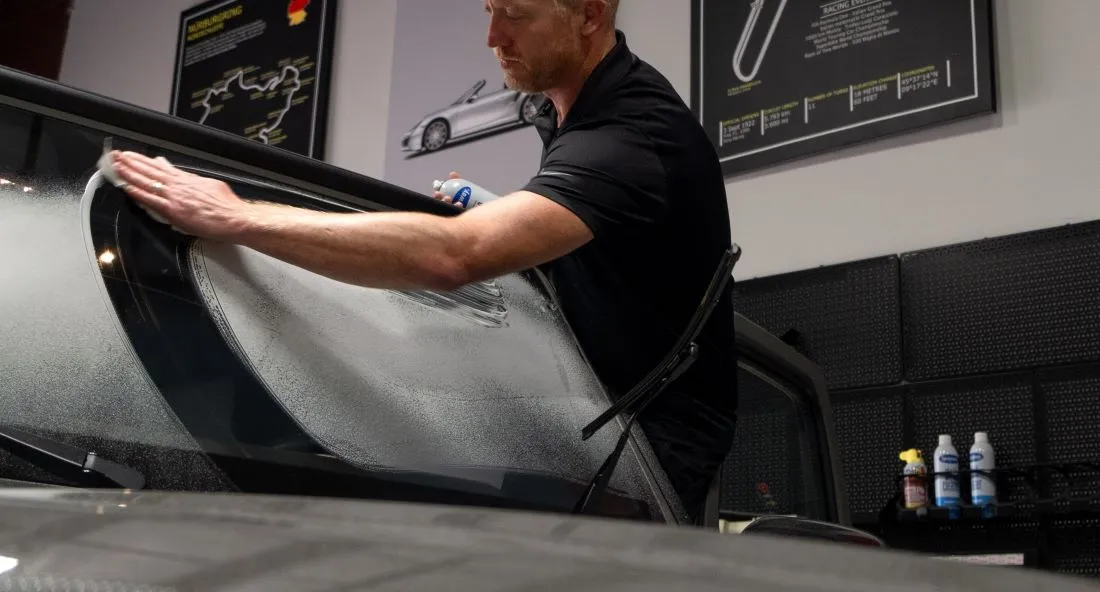 A windshield with glass cleaner sprayed on it and a man holding a can of glass cleaner in one hand and cleaning the windshield with the other