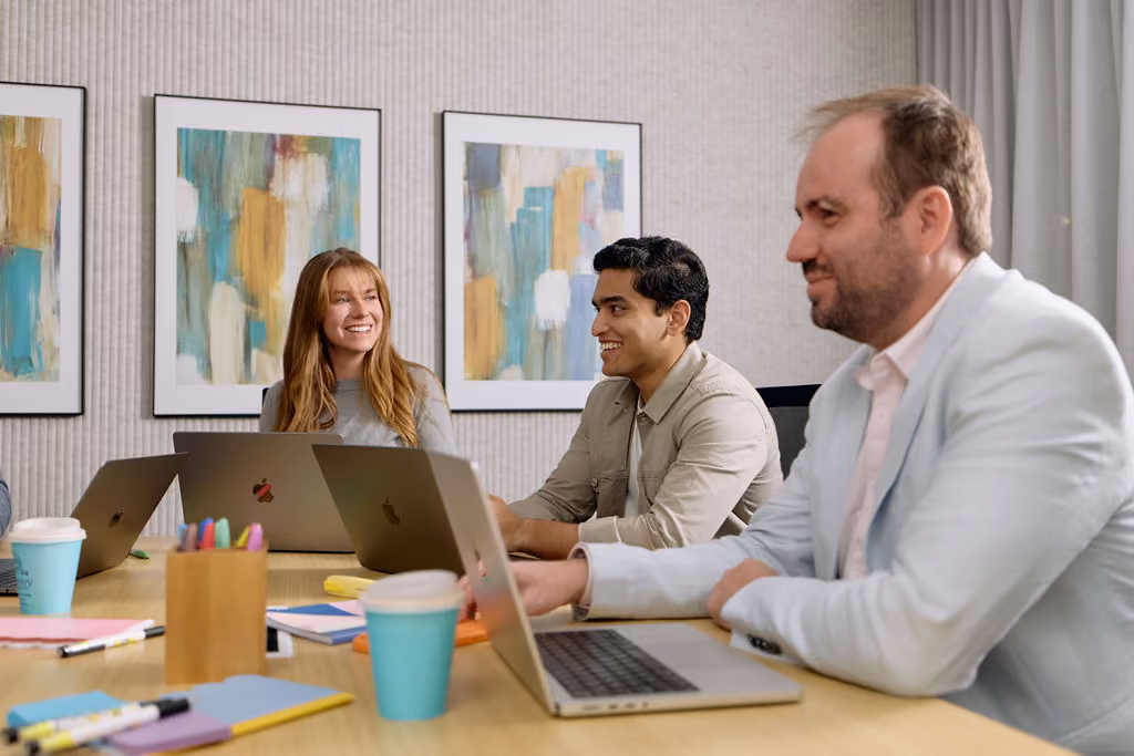 Three colleagues seated at a table with laptops, working together in a bright meeting space, from Agend's day-in-the-life content shoot.