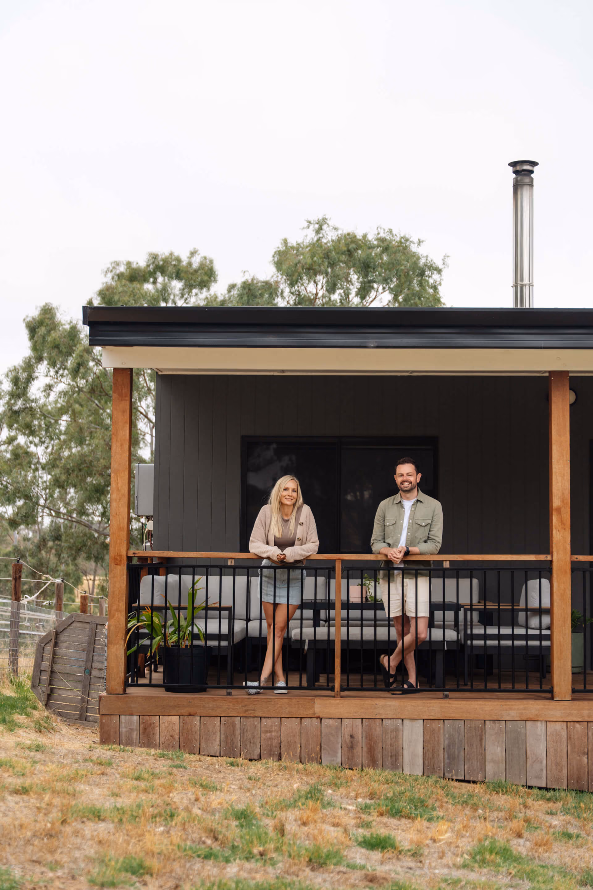Two people leaning on the balustrade of their timber veranda of a Modify home in the Adelaide Hills, with a chimney flue visible on the rooftop.
