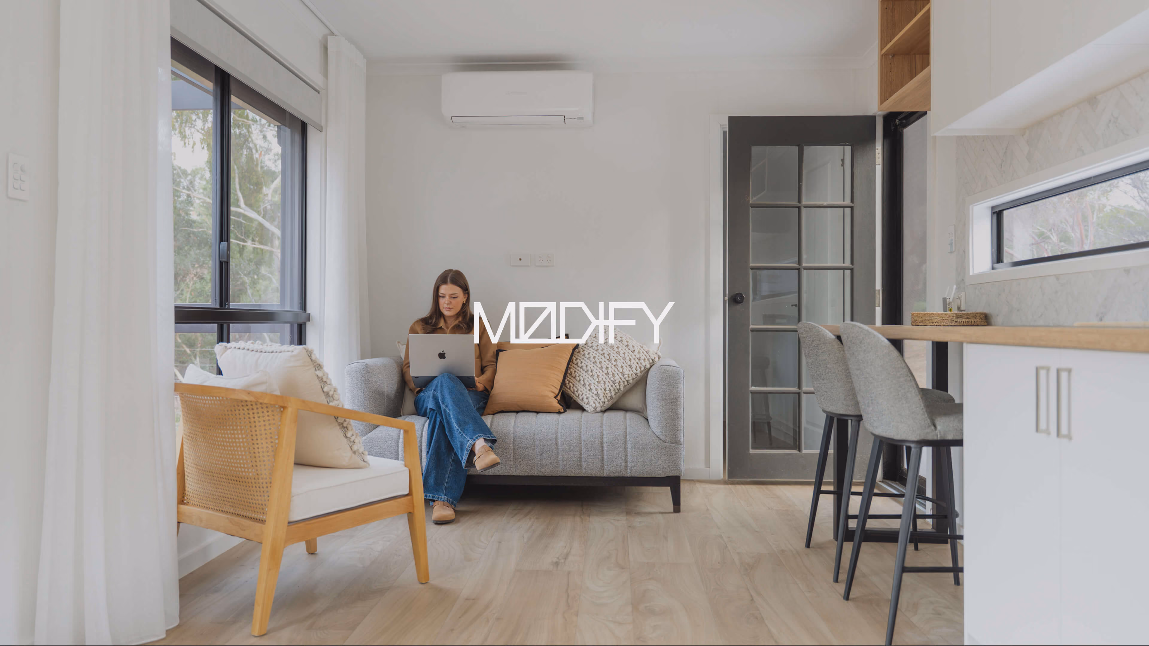 Woman relaxing with a laptop in the bright, modern living space of a Modify Modular home, with the Modify logo overlaid.