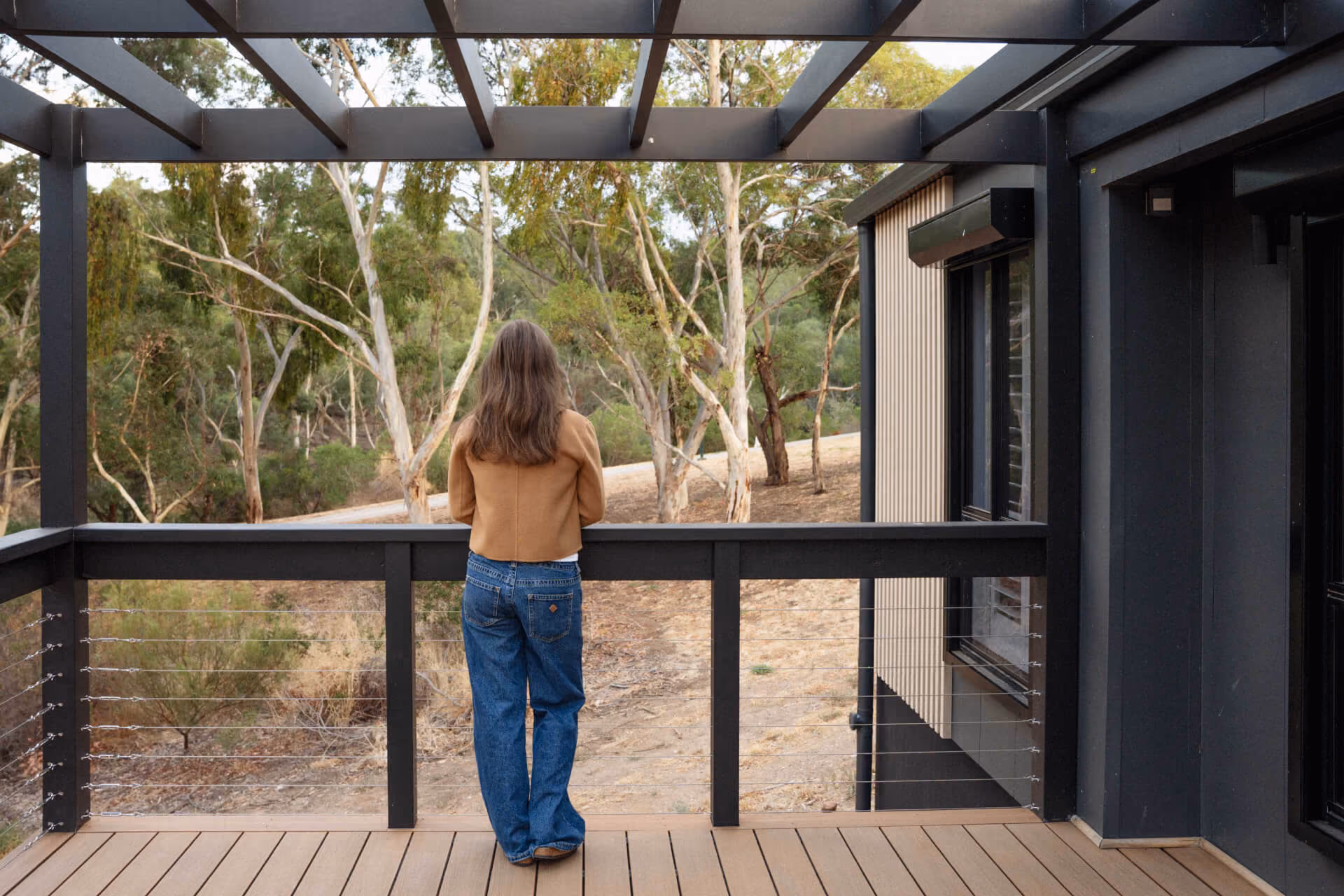 Woman standing on the timber front deck under a pergola at a Modify modular home in Paradise, Adelaide, looking out toward surrounding native gum trees.