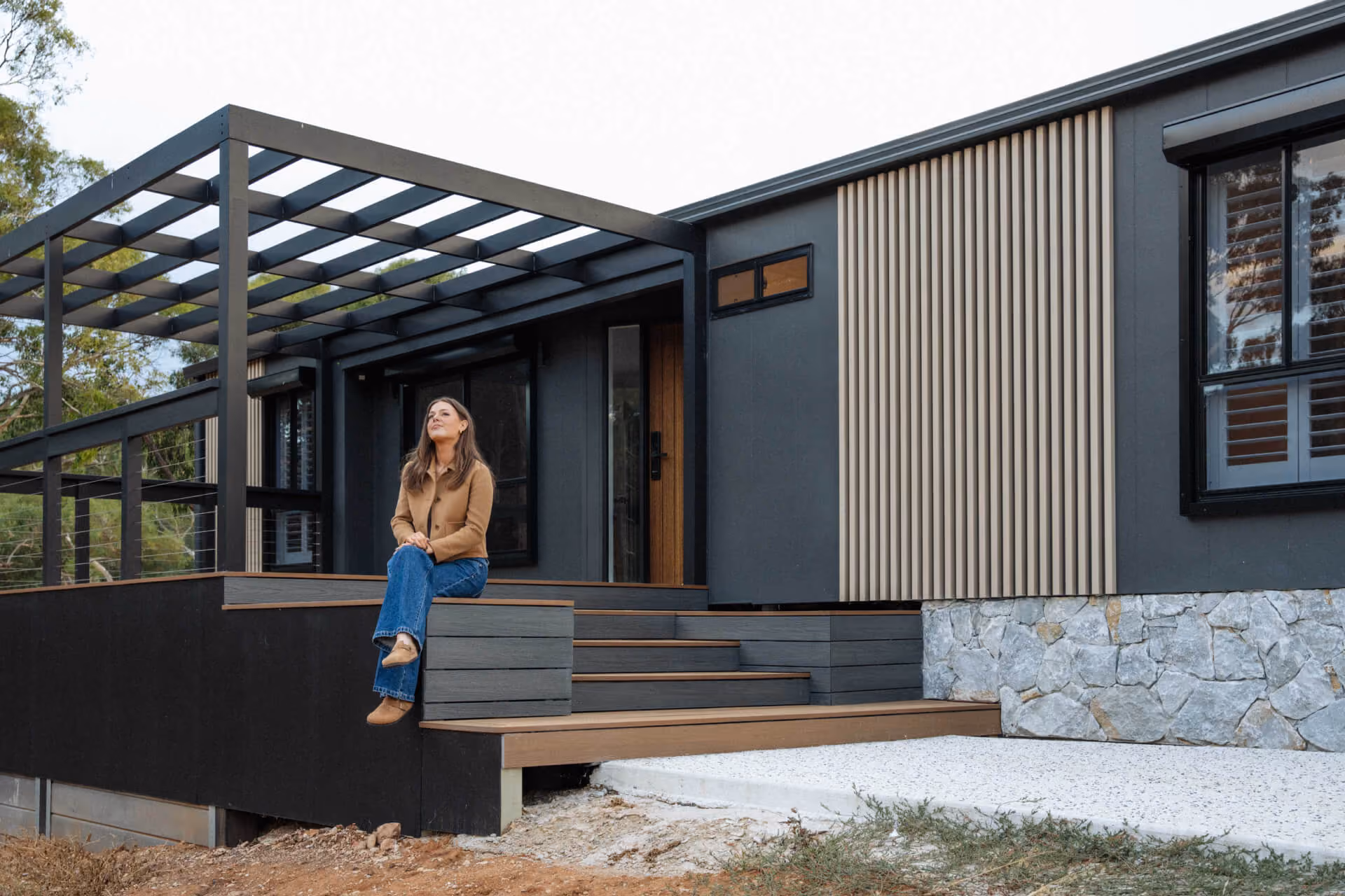 Wide exterior view of a Modify portable home in Paradise, Adelaide, showing the front entrance with pergola, mixed timber and steel cladding, and a woman seated on the front steps.