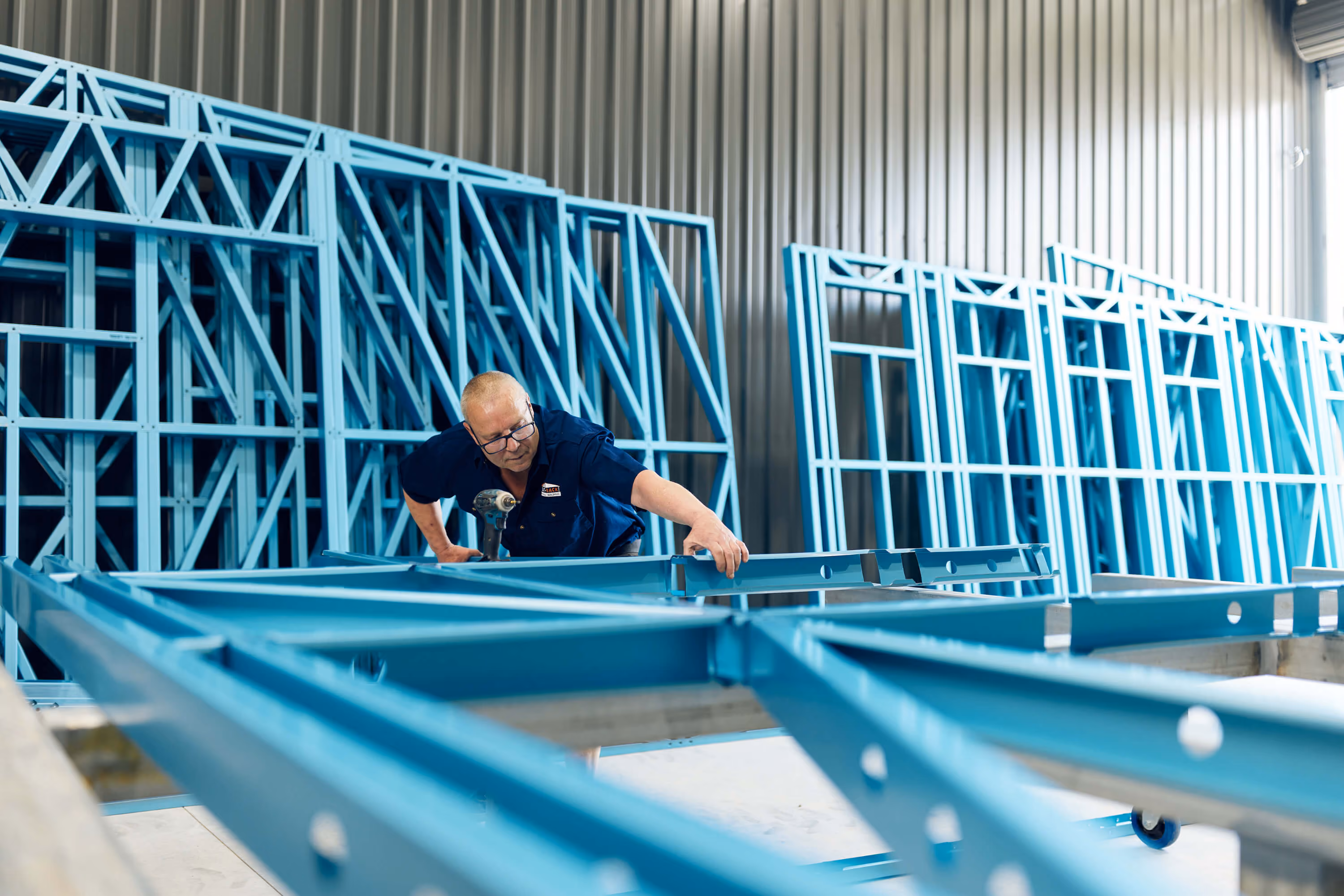 Modify Steel worker assembling a blue steel frame in the workshop, with finished frames lined up behind him. 