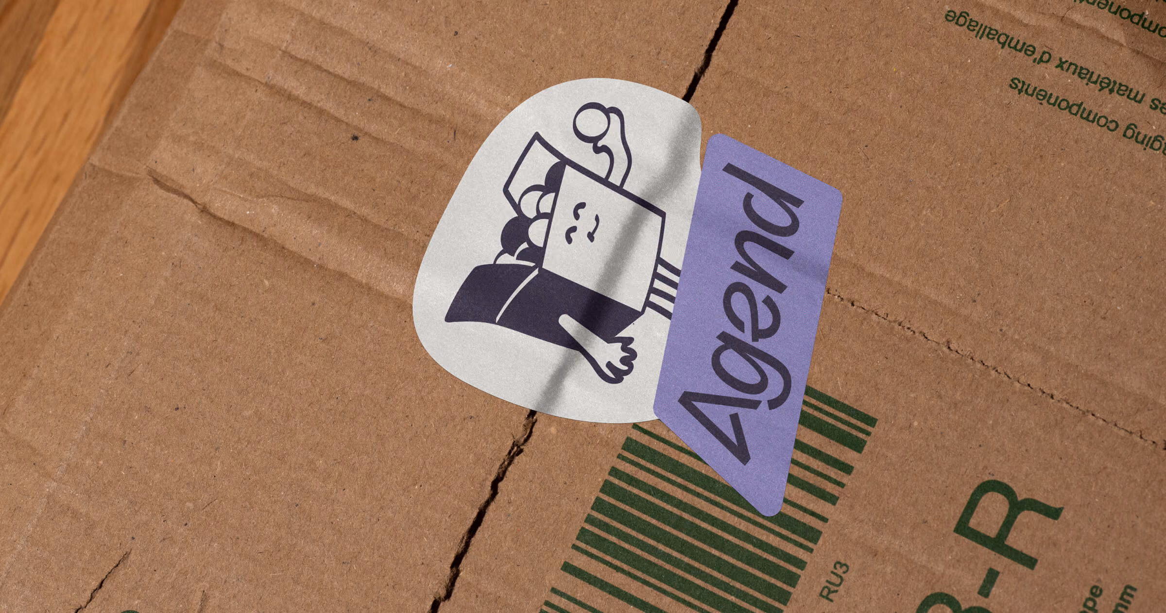 Agend’s box mascot sticker and purple logo label on a cardboard shipping box, bringing playful brand personality to everyday packaging.